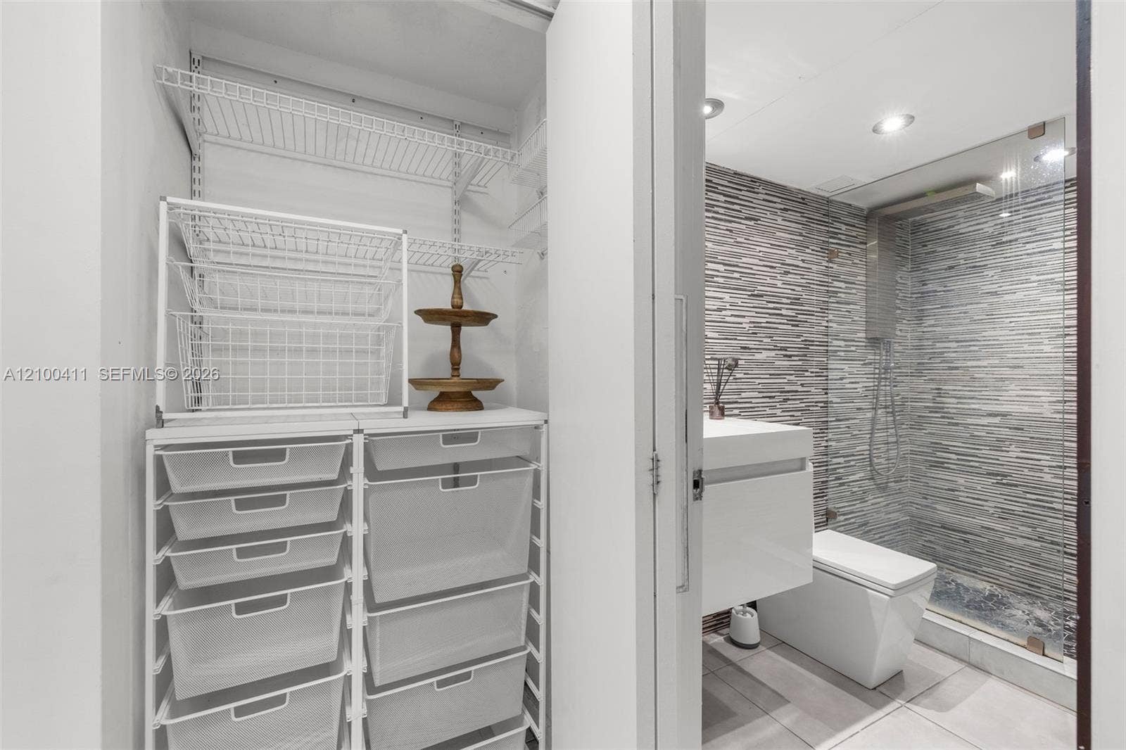 Walk-in closet featuring wire shelving and a modular storage system, paired with a bathroom containing a wall-mounted vanity, square-profile toilet, glass-enclosed shower with horizontal textured tile, and recessed lighting