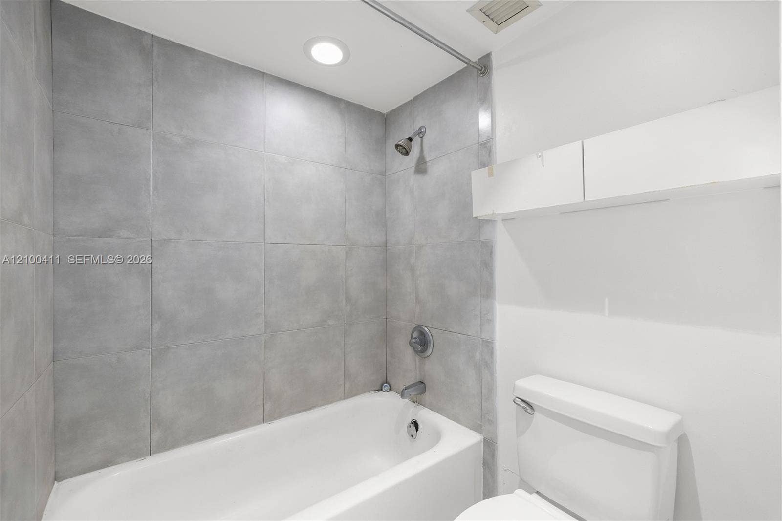 Full-height grey tile tub surround, white porcelain bathtub, wall-mounted shower head, recessed ceiling lighting, and built-in white upper cabinetry