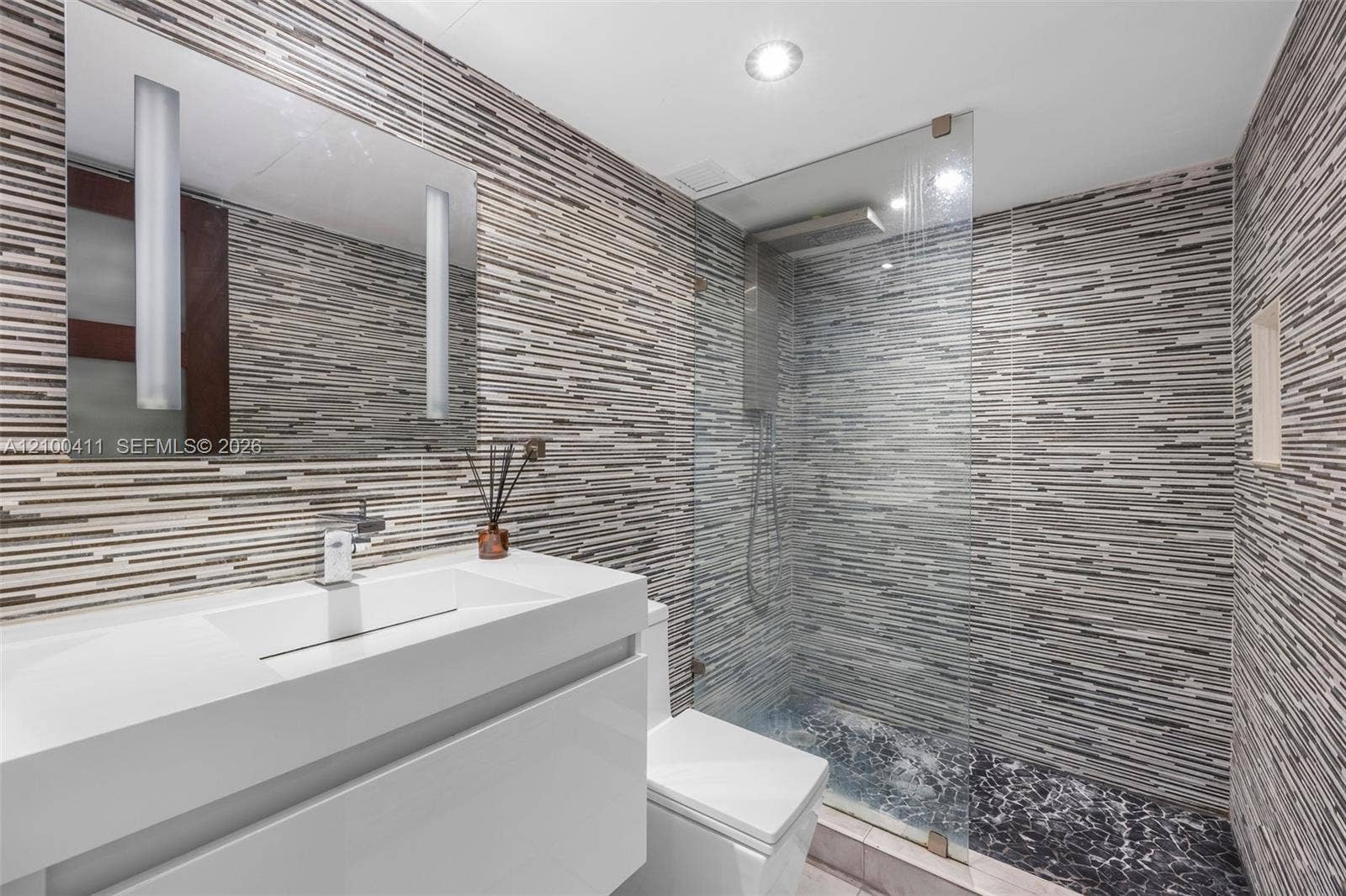 Modern floating vanity with a seamless integrated sink, wall-mounted faucet, linear multi-toned tile wall covering, frameless glass shower enclosure, and recessed shower shelving