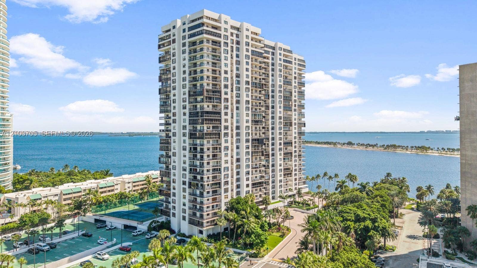 High-rise residential tower featuring private balconies, tennis courts, covered parking, and palm tree landscaping situated along the waterfront