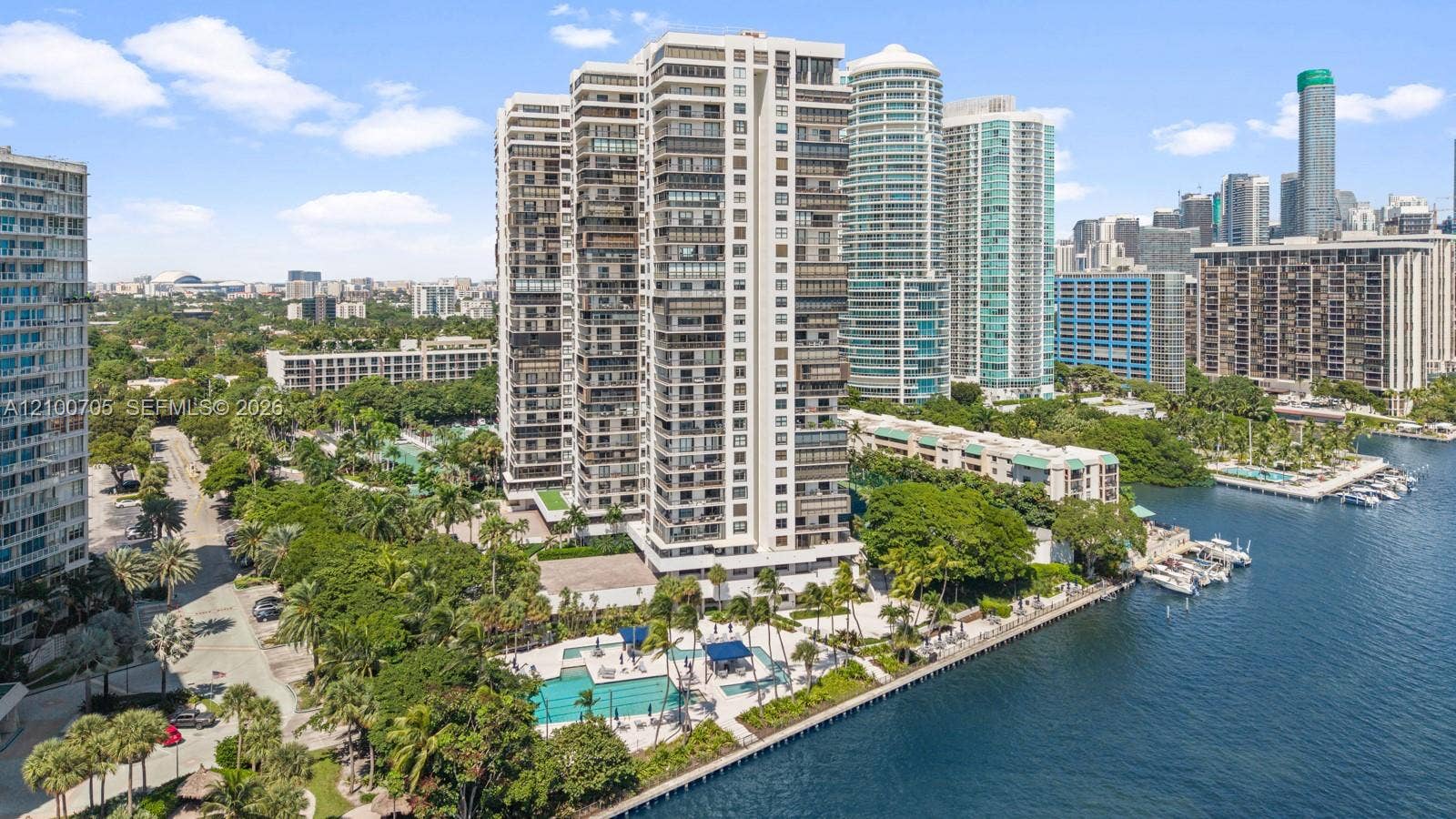 High-rise residential building featuring floor-to-ceiling glass paneling, a ground-level swimming pool, expansive waterfront frontage, and mature landscaping