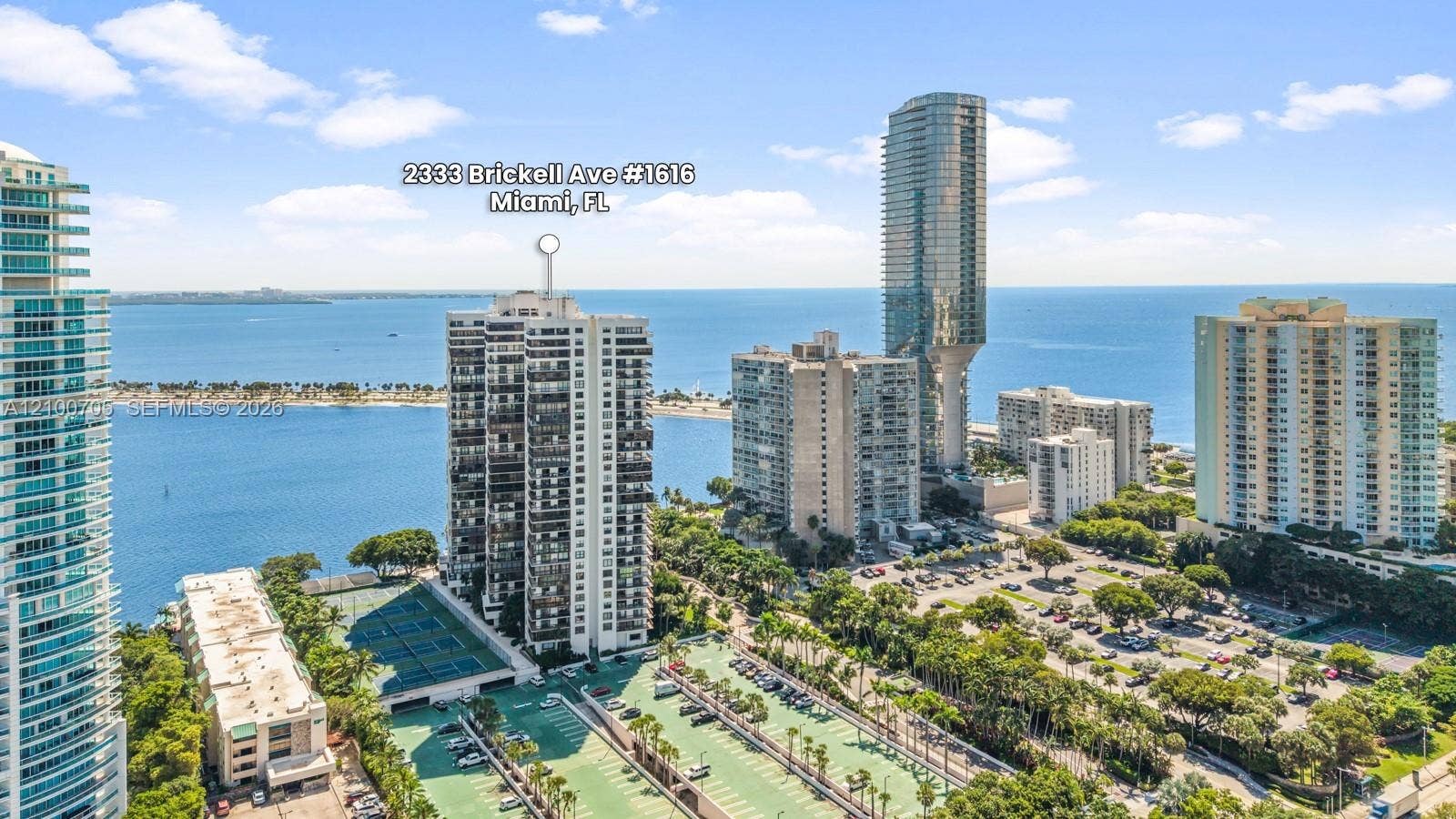 High-rise residential building featuring expansive water frontage, deep balconies, and tennis courts situated within a landscaped grounds setting