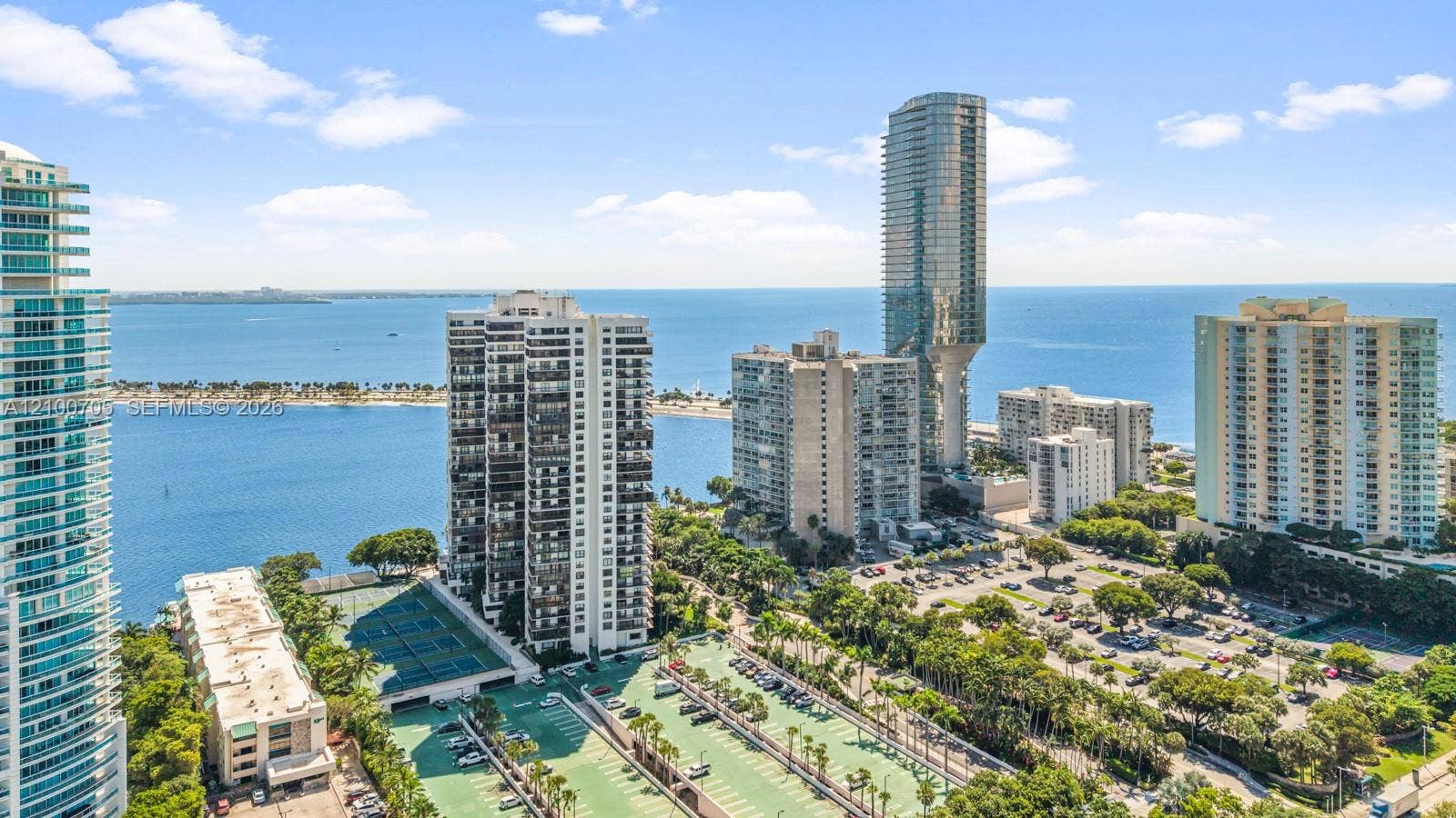 High-rise coastal cityscape featuring expansive water frontage, proximity to public tennis courts, mature tropical landscaping, and integrated surface parking