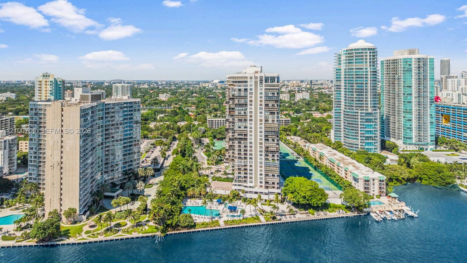 Aerial perspective featuring high-rise architecture, waterfront bulkhead, swimming pools, tennis courts, and landscaped grounds