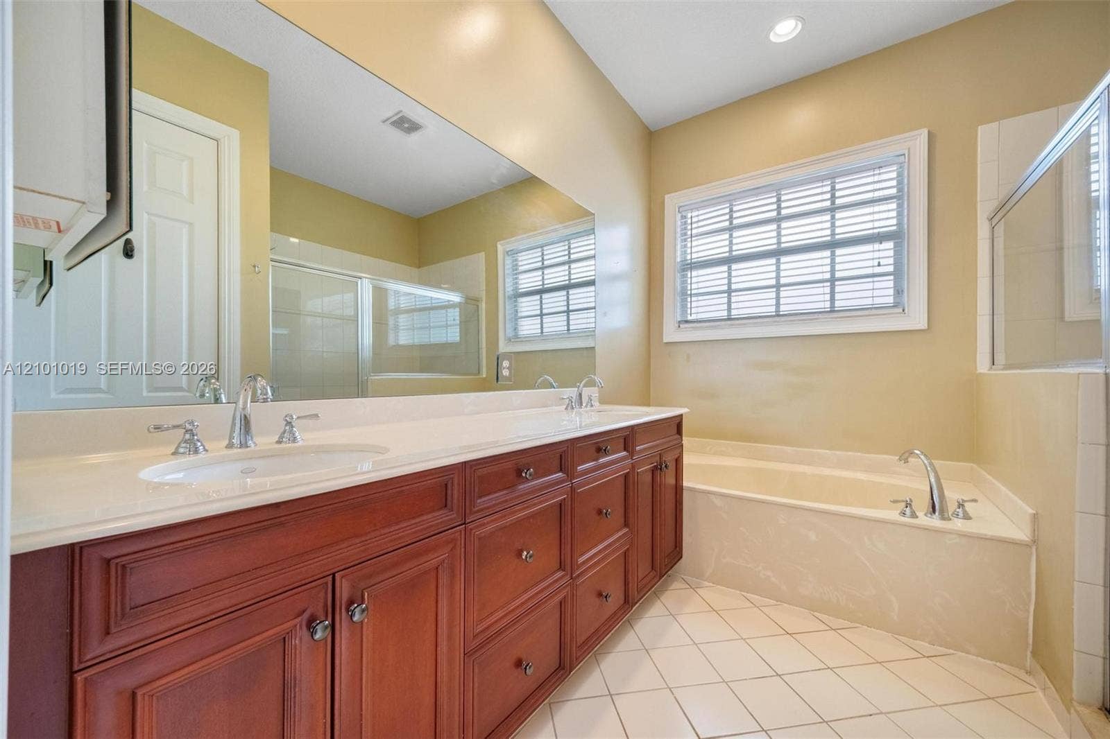 Extended wood-finish cabinetry featuring dual vanity sinks, expansive wall-to-wall mirror, built-in soaking tub with surround, and tile flooring