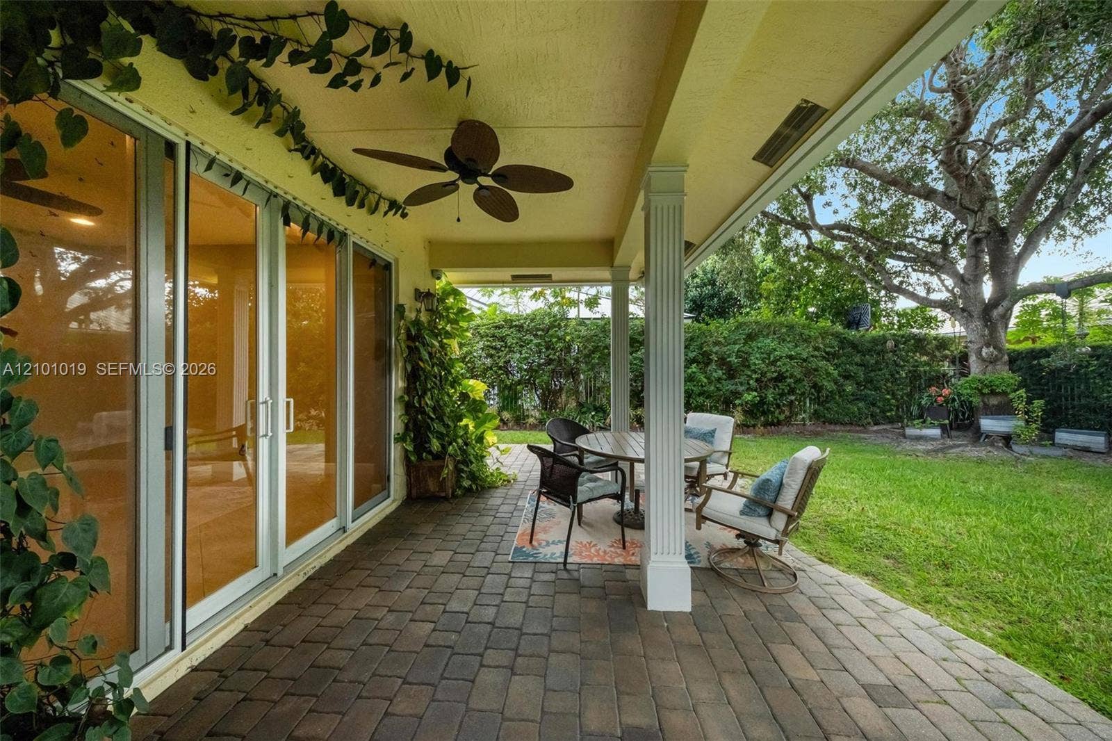 Covered patio with brick-style paver flooring, fluted columns, ceiling fan, and multi-panel sliding glass doors