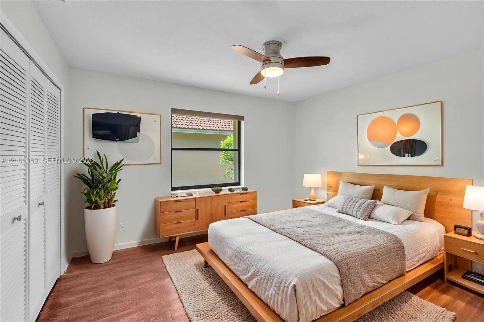 Bedroom featuring wood-finish flooring, white louvered closet doors, a central ceiling fan with integrated lighting, and a window with a neutral roller shade -virtually staged