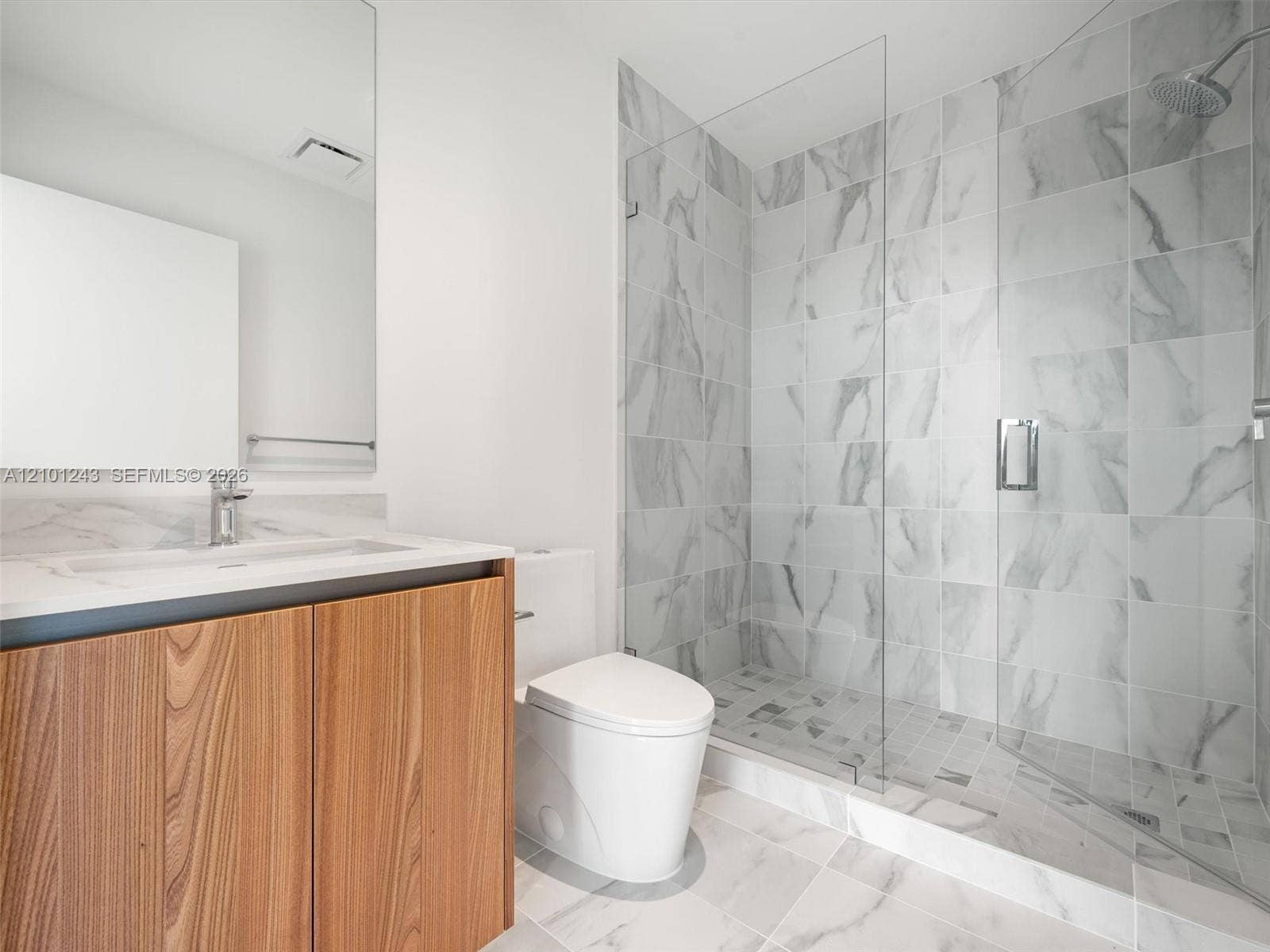 Bathroom featuring a wood-finish vanity with a stone-surfaced countertop, large-format tile flooring, walk-in glass-enclosed shower with floor-to-ceiling tile surround, and a wall-mounted mirror