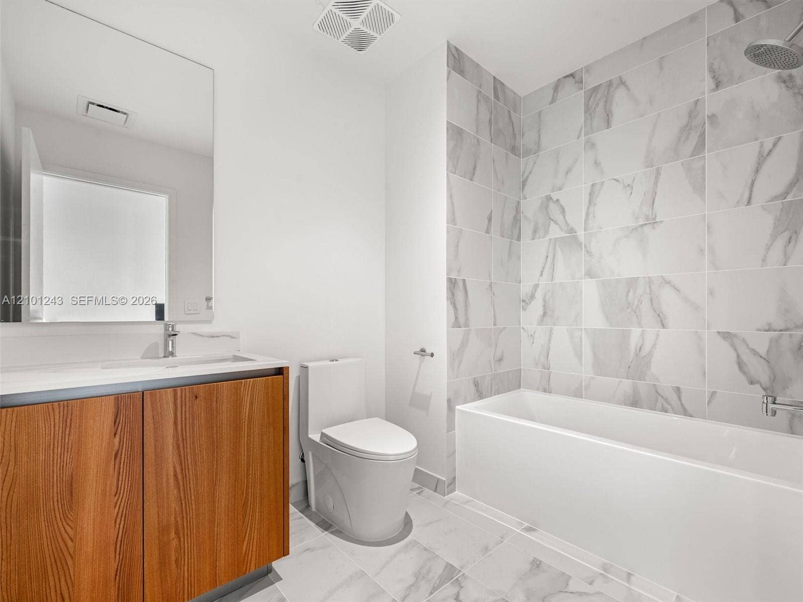 Bathroom featuring a wood-finish vanity with a rectangular sink, large frameless mirror, white plumbing fixtures, and floor-to-ceiling tile surrounding the bathtub