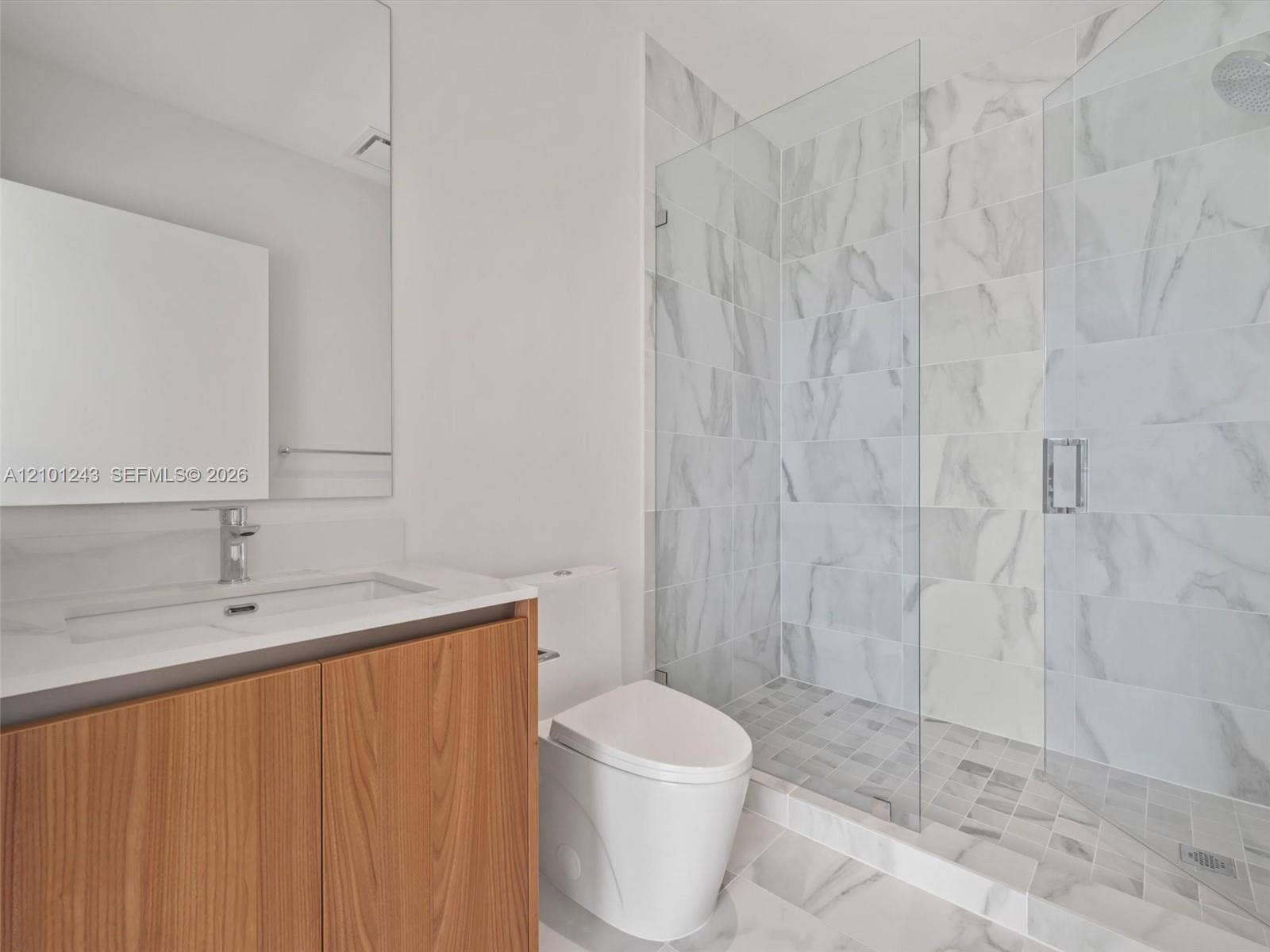 Bathroom featuring a vanity with a light-toned wood-finish cabinet and solid-surface countertop, a frameless glass-enclosed shower with large-format wall tile, and a wall-mounted mirror