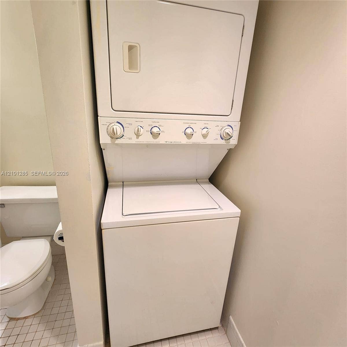 Stacked white laundry center featuring top-load washer and dryer configuration, paired with a white porcelain toilet on tiled flooring