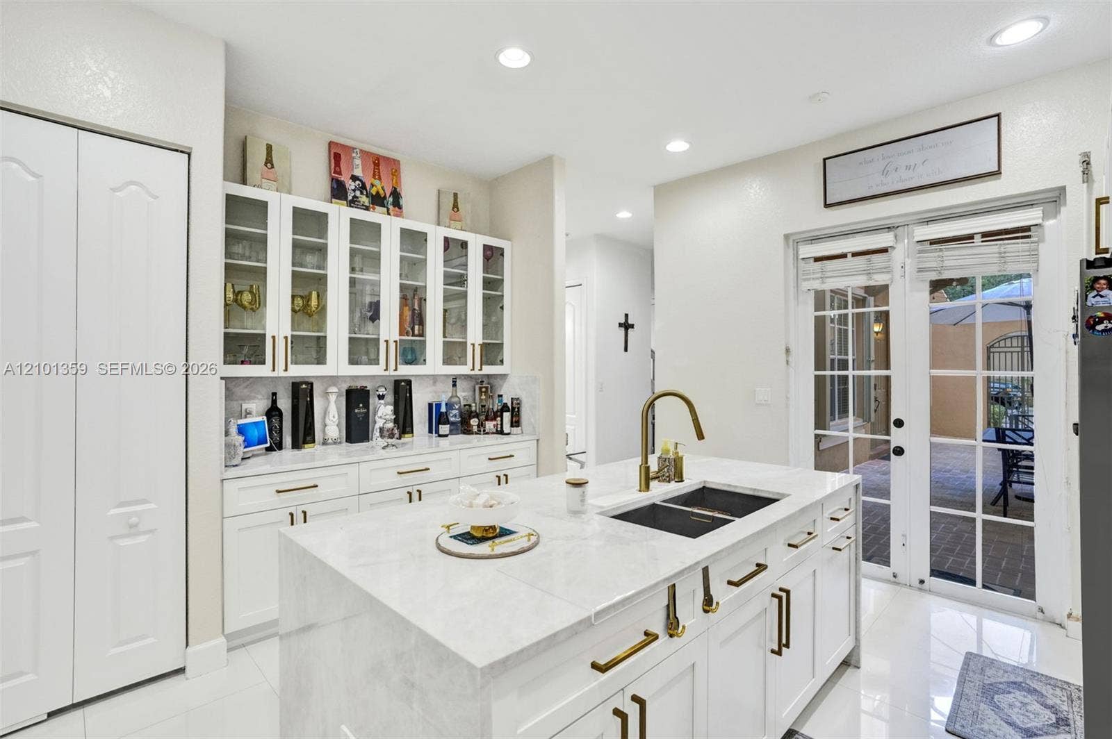 859 SW 147th Terrace, #0, Pembroke Pines, FL 33027 Kitchen island featuring a stone countertop, an undermount dual-basin sink, and a brass-finish faucet, complemented by built-in white cabinetry with glass-front display uppers, brass-finish hardware, and French doors