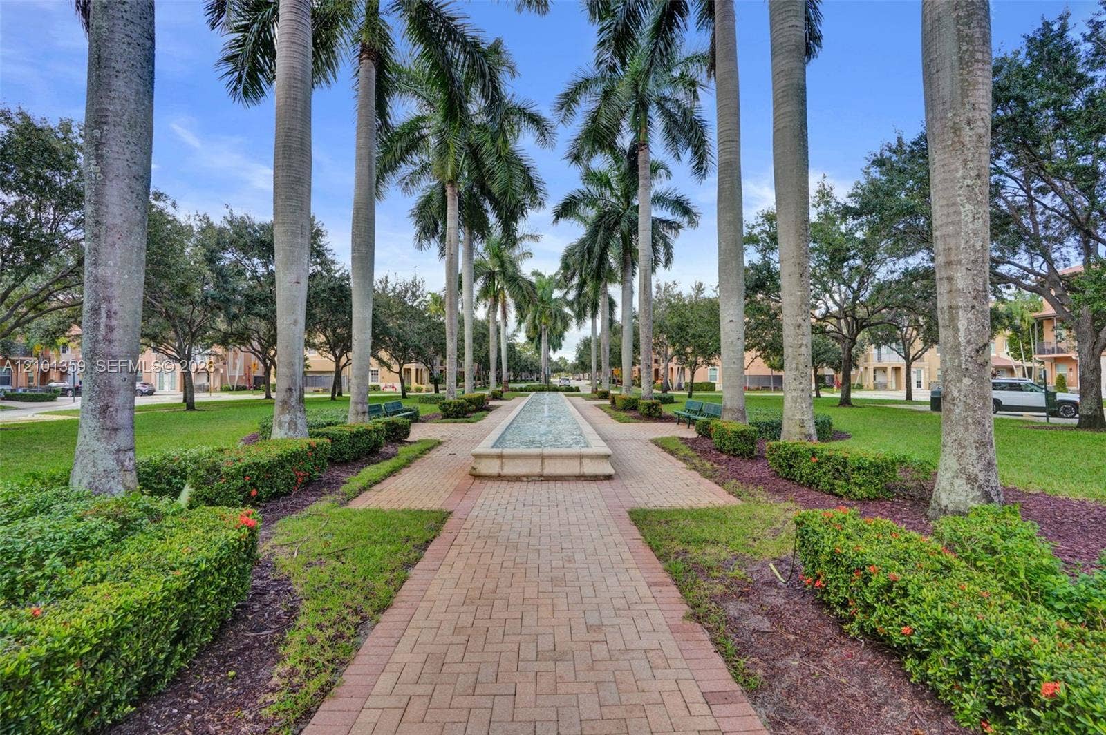 859 SW 147th Terrace, #0, Pembroke Pines, FL 33027 Landscaped grounds featuring a central stone-clad fountain, brick-paved walkway, mature palm trees, manicured hedges, and expansive lawn areas