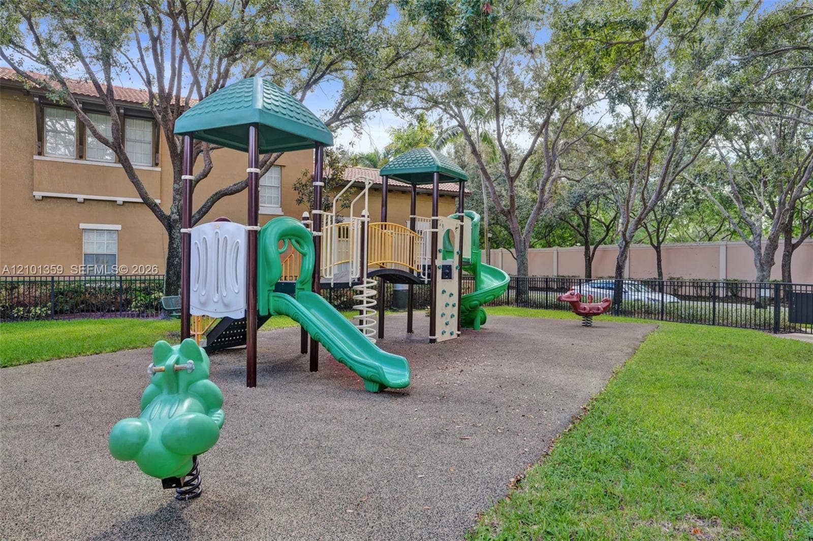 859 SW 147th Terrace, #0, Pembroke Pines, FL 33027 Recreational area featuring a multi-component play structure with slides and climbing elements, set within a landscaped perimeter enclosed by metal fencing and mature shade trees