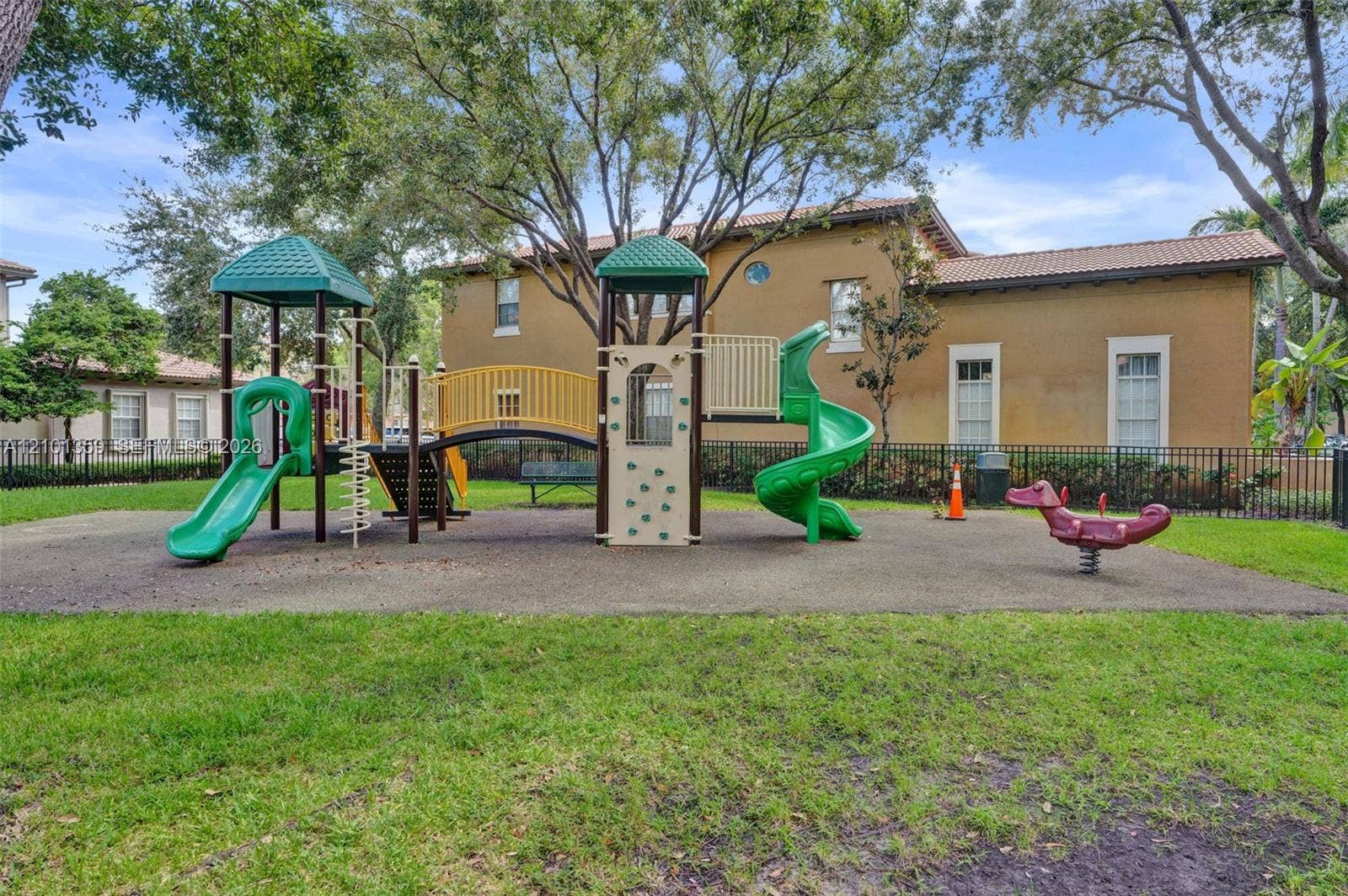 859 SW 147th Terrace, #0, Pembroke Pines, FL 33027 Community playground featuring a multi-level structure with two slides, a bridge, climbing elements, and a spring rider set within a grass-covered area enclosed by black metal fencing