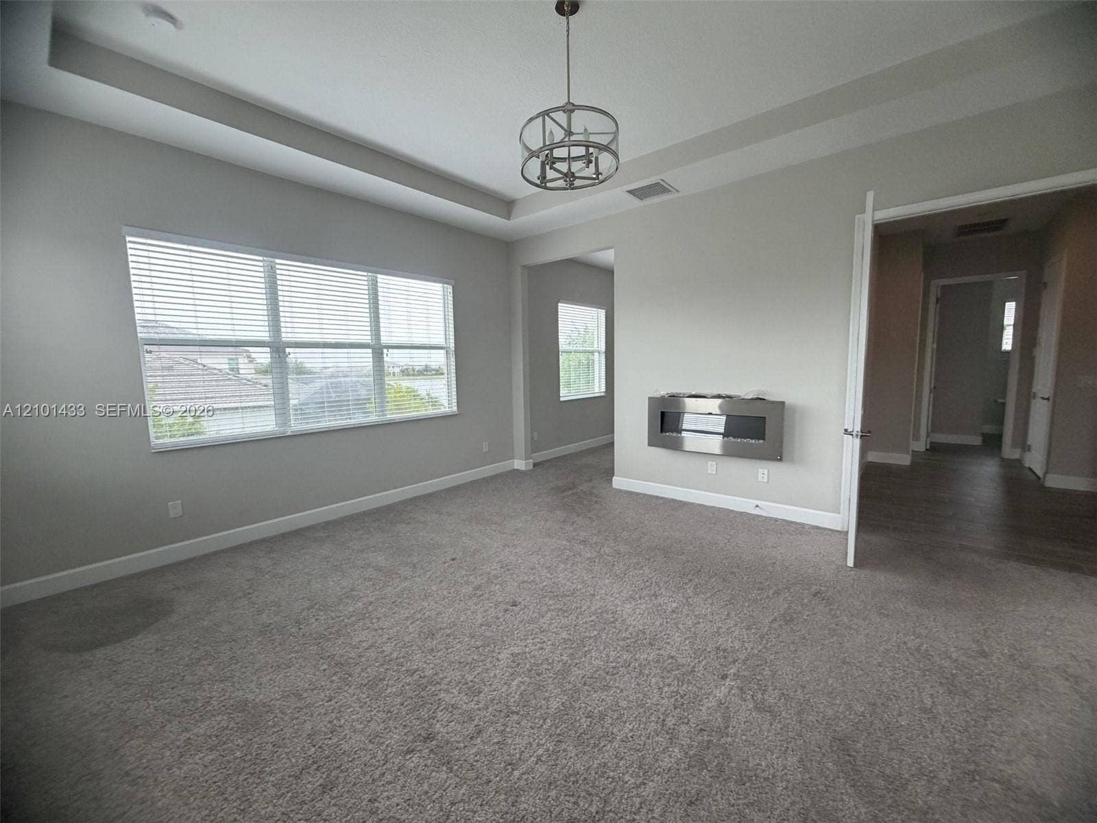 Tray ceiling with a modern chandelier, wall-mounted electric fireplace, and light-colored carpeting