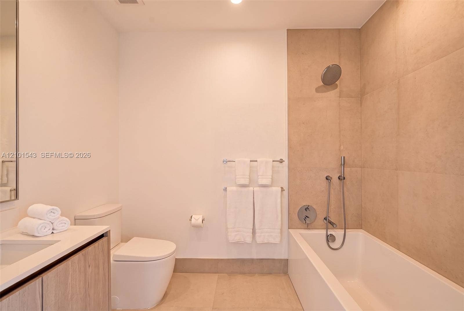 Bathroom featuring a wood-finish vanity, large-format neutral wall tile, integrated tub and shower combination with handheld fixture, and recessed lighting