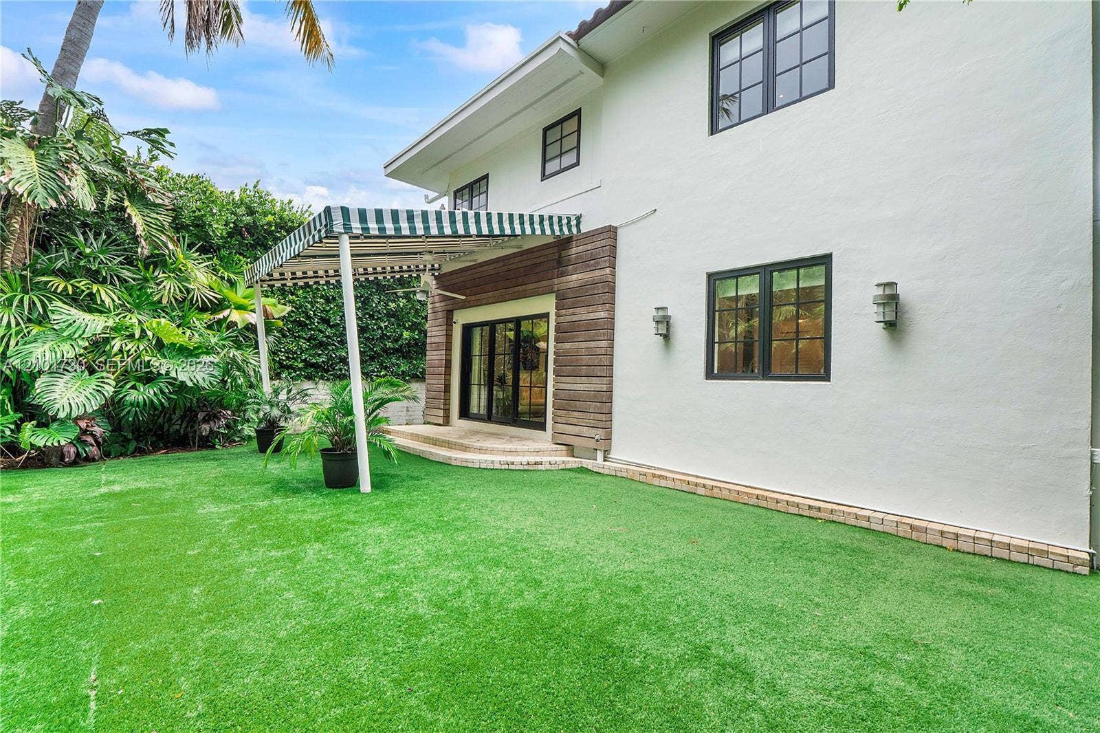 435 W 51st Street, Miami Beach, FL 33140 Exterior space featuring a manicured lawn, tiered stone patio, attached striped awning, wood-slat siding accents, and multi-pane windows