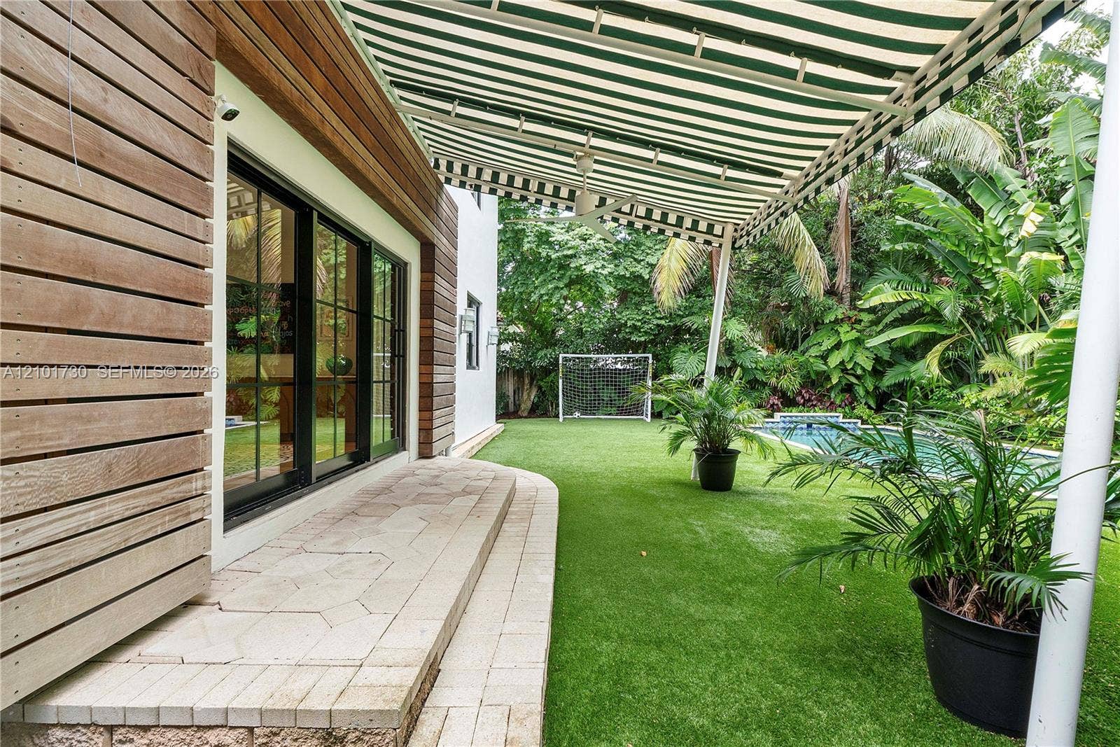 435 W 51st Street, Miami Beach, FL 33140 Exterior features include horizontal wood-slat siding, a retractable striped awning, a stone-paver patio, a swimming pool, and artificial turf lawn
