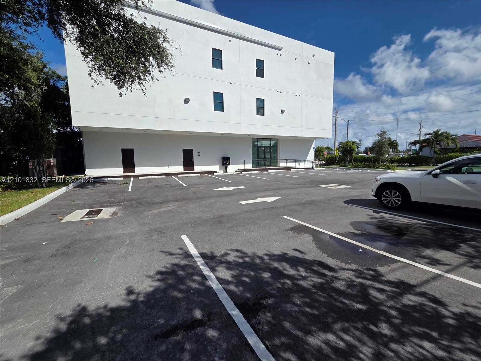 401 Pembroke Road, #201, Hallandale Beach, Hallandale Beach, FL 33009 Three-story white stucco exterior featuring a paved asphalt parking lot with designated spaces, dual entry doors, and a glass-paneled entrance with an integrated ramp