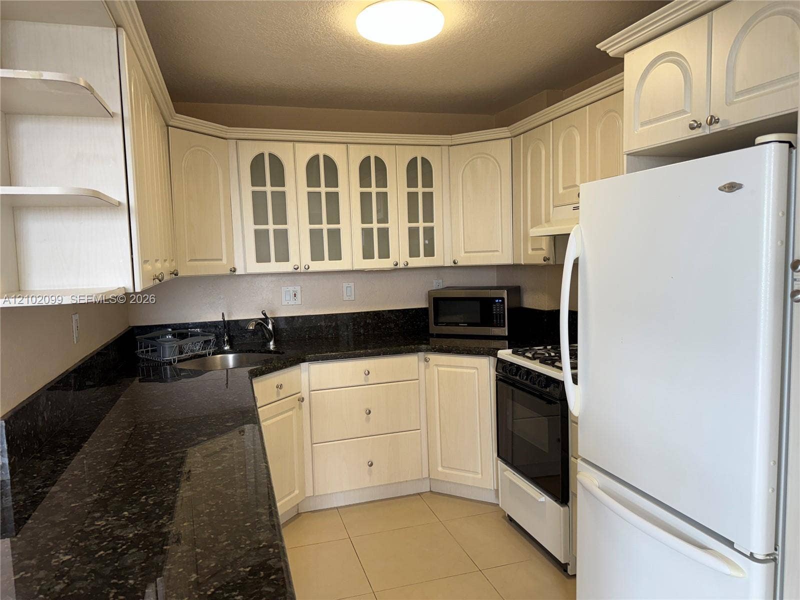 13120 SW 92nd Avenue, #B-PH1, Miami, Miami, FL 33176 Kitchen featuring light-toned cabinetry with glass-panel upper doors, dark stone-finish countertops, tile flooring, and integrated white appliances