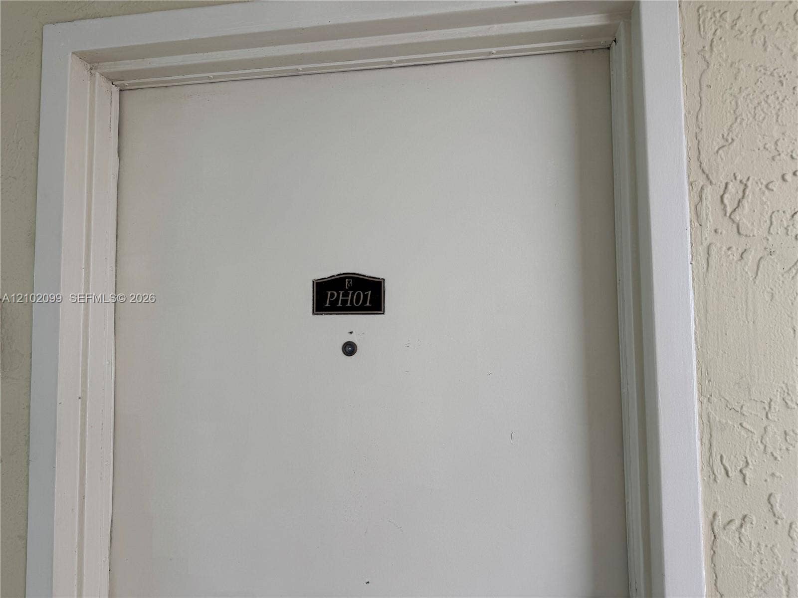 13120 SW 92nd Avenue, #B-PH1, Miami, Miami, FL 33176 Solid-core white entry door featuring an arched metal identification plaque and a built-in security peephole, set within a neutral-toned textured wall