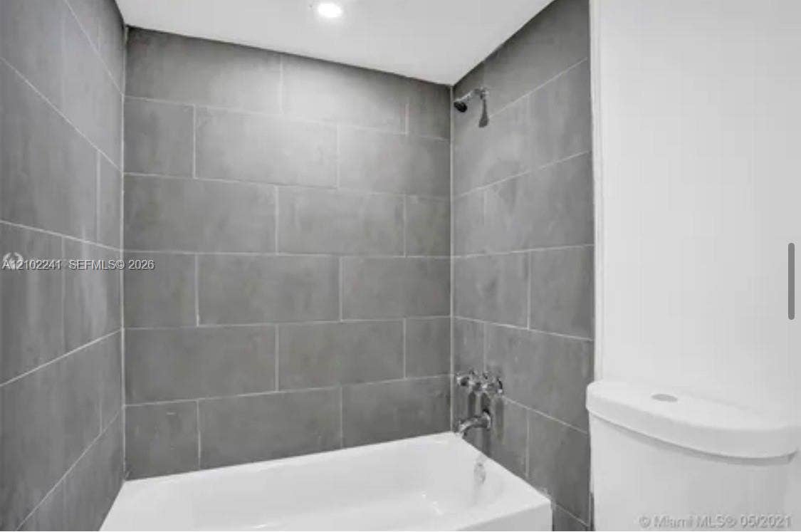Full-height gray ceramic wall tiling, white porcelain bathtub, chrome wall-mounted shower head, chrome faucet fixtures, and white toilet