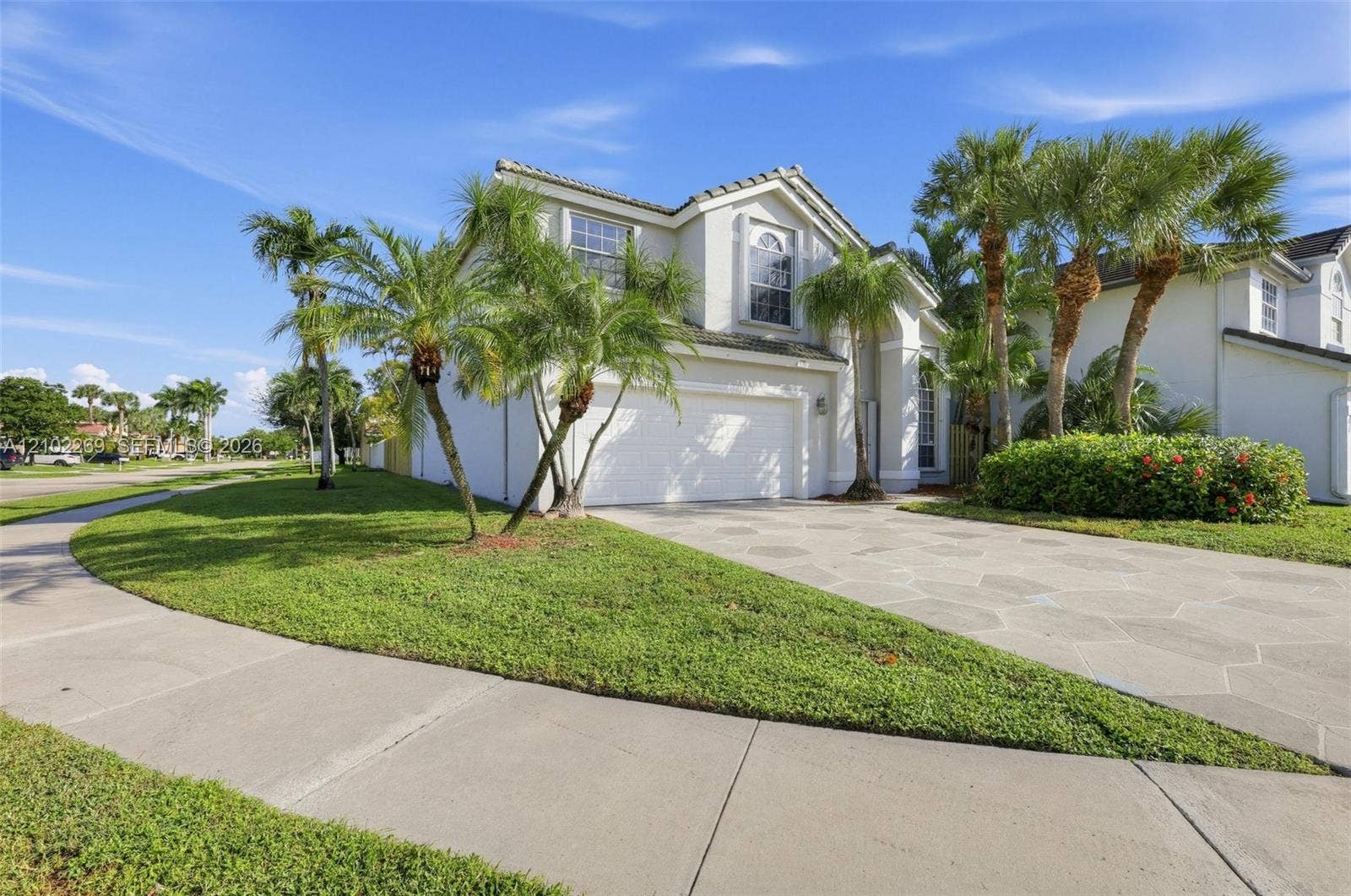 Two-story stucco exterior featuring a multi-gabled roofline, expansive concrete paver driveway, attached two-car garage, and mature palm landscaping