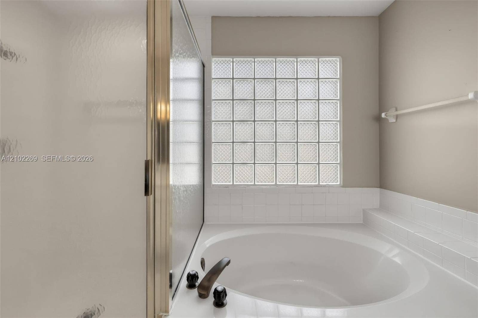 Oval soaking tub with a white tile surround, glass block window, and metal-finished hardware