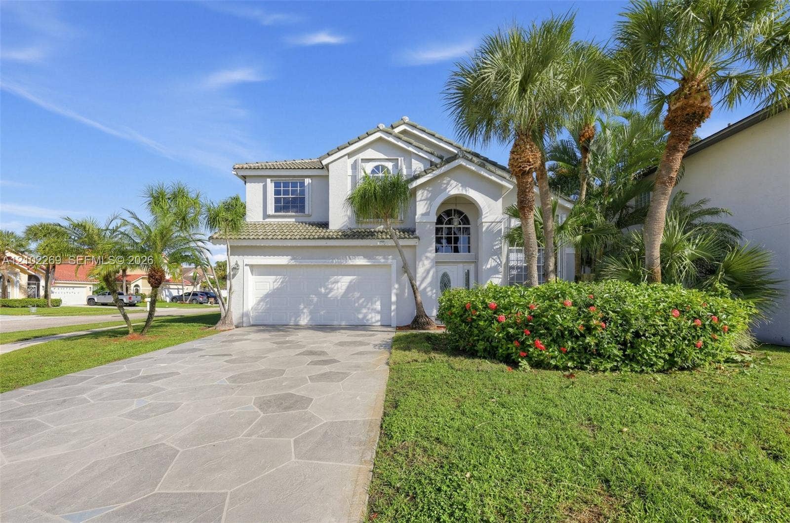Two-story stucco exterior featuring a tiled roof, arched entryway, polygon-patterned paver driveway, and established palm landscaping