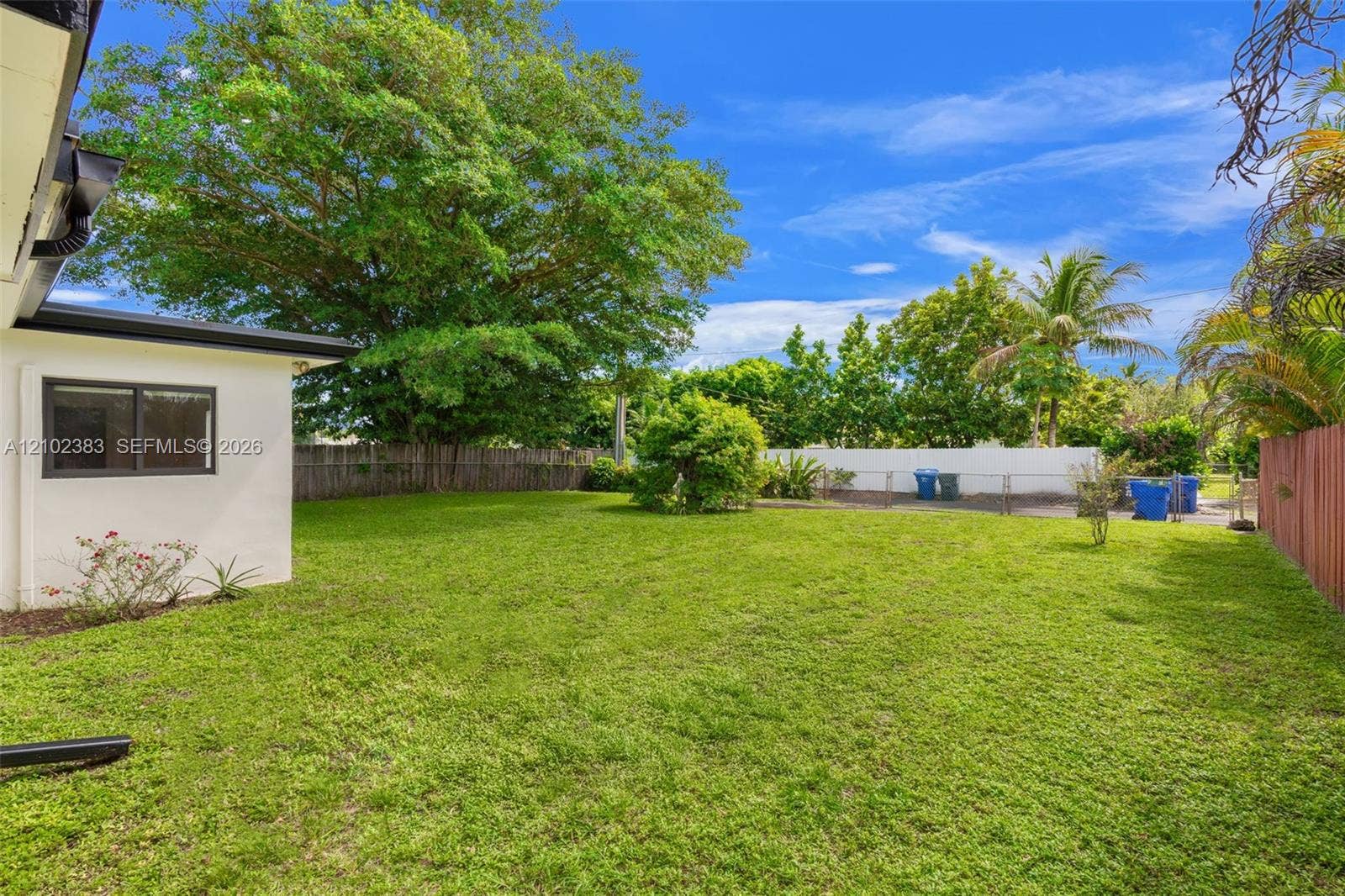 Expansive backyard featuring a manicured lawn, mature trees, white vinyl privacy fencing, and a side wood-panel fence