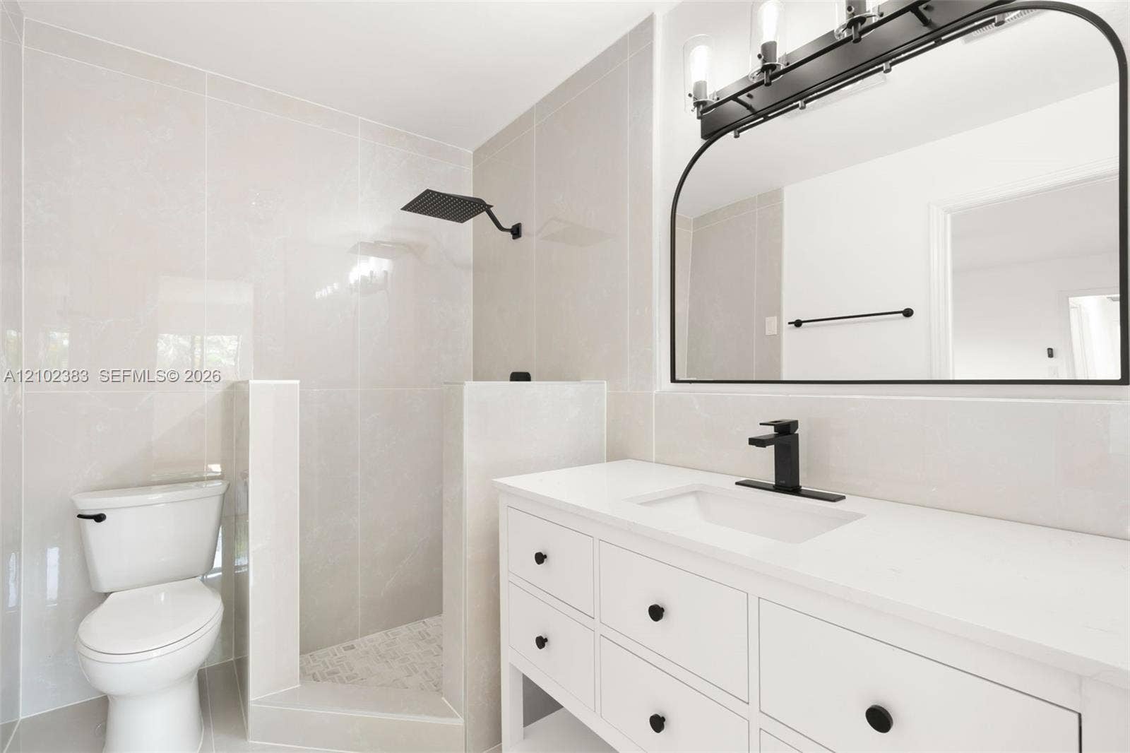 Primary bathroom featuring a white vanity with a quartz countertop, matte black faucet, wall-mounted rain shower head, arched vanity mirror, and light-toned wall tile