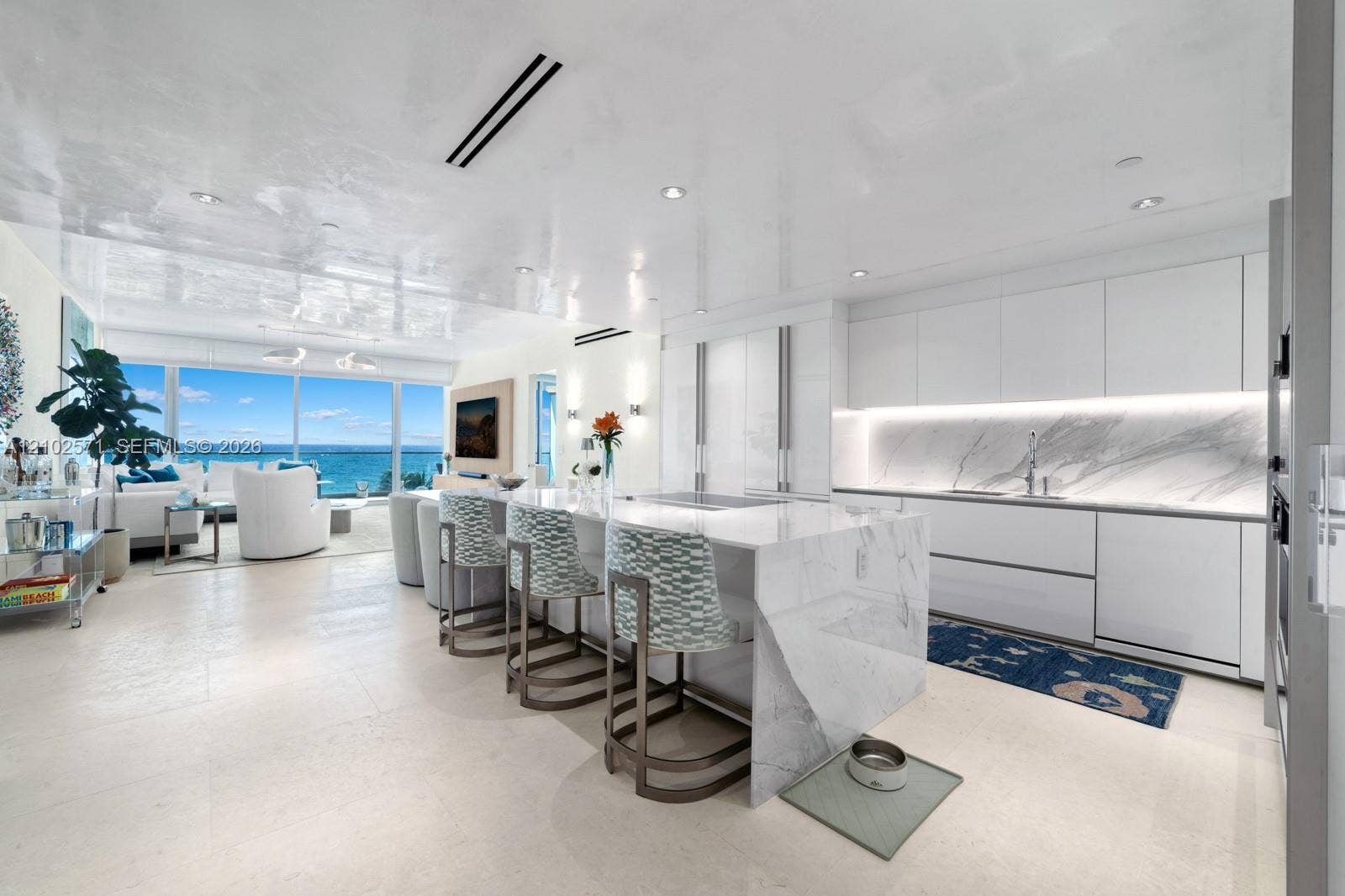 10201 Collins Avenue, #502, Bal Harbour, FL 33154 Contemporary kitchen featuring a waterfall-edge island, stone slab backsplash, flat-panel cabinetry, and integrated lighting, set against floor-to-ceiling windows