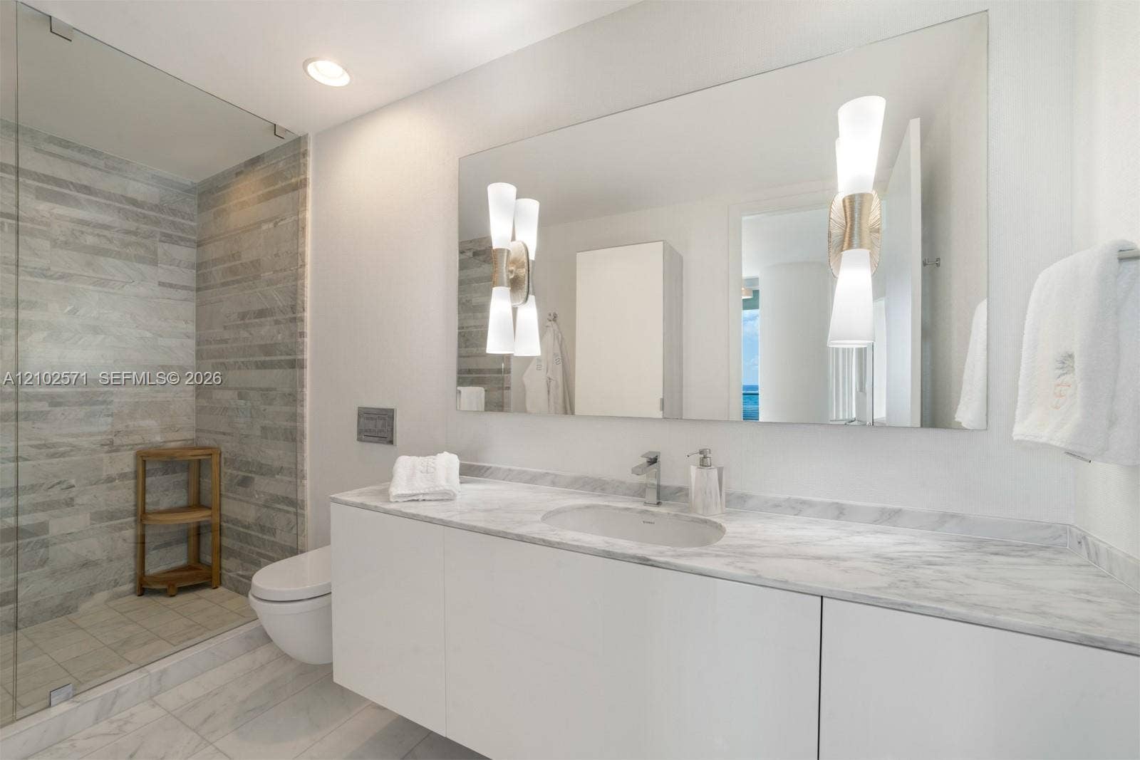 10201 Collins Avenue, #502, Bal Harbour, FL 33154 Floating vanity with integrated sink, stone-surface countertop, walk-in glass-enclosed shower featuring horizontal stone-tiled walls, wall-mounted toilet, and dual decorative light fixtures
