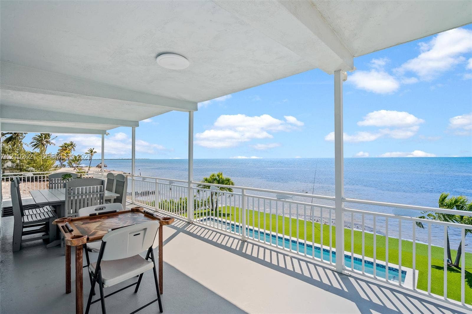 Covered exterior balcony with a white railing, overlooking an in-ground swimming pool and expansive body of water