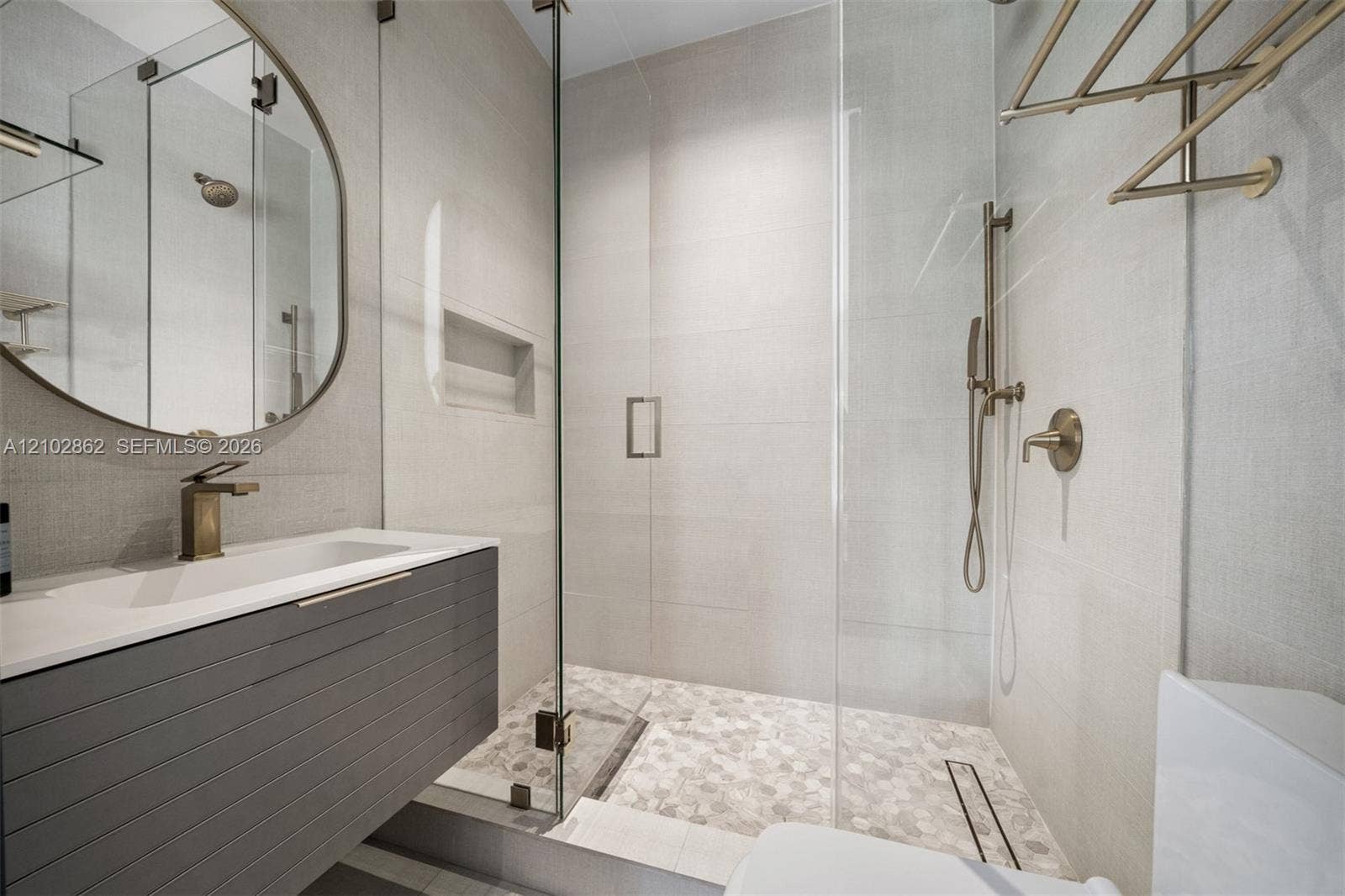 Floating vanity with a smooth top, glass-enclosed shower featuring a pebble-tiled floor, integrated wall niche, brass-finish fixtures, and a circular wall-mounted mirror