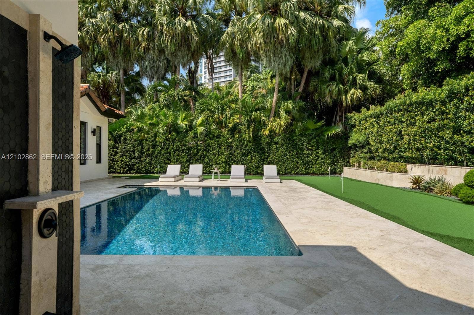 Outdoor pool area featuring natural stone decking, a synthetic turf putting green, a privacy hedge, and a wall-mounted outdoor shower with dark stone surround