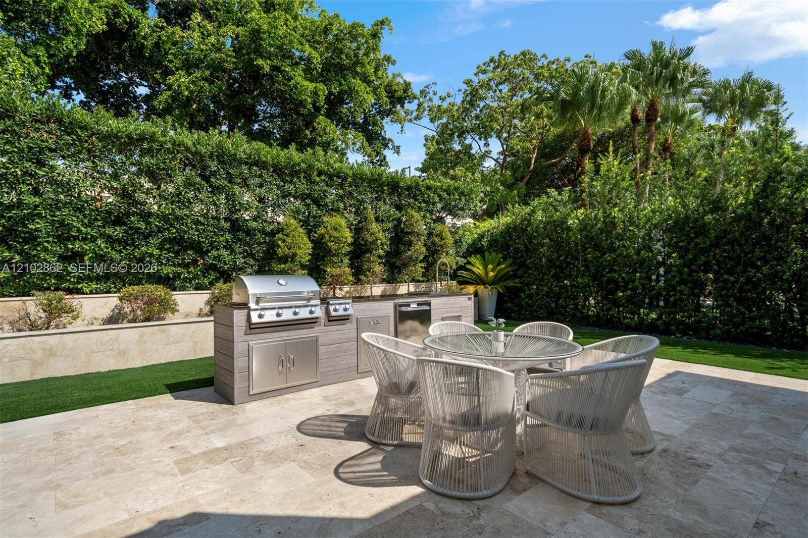 Outdoor kitchen featuring a stainless steel grill, a built-in refrigerator, stone-finish countertops, and a stone-tiled patio bordered by mature privacy hedges