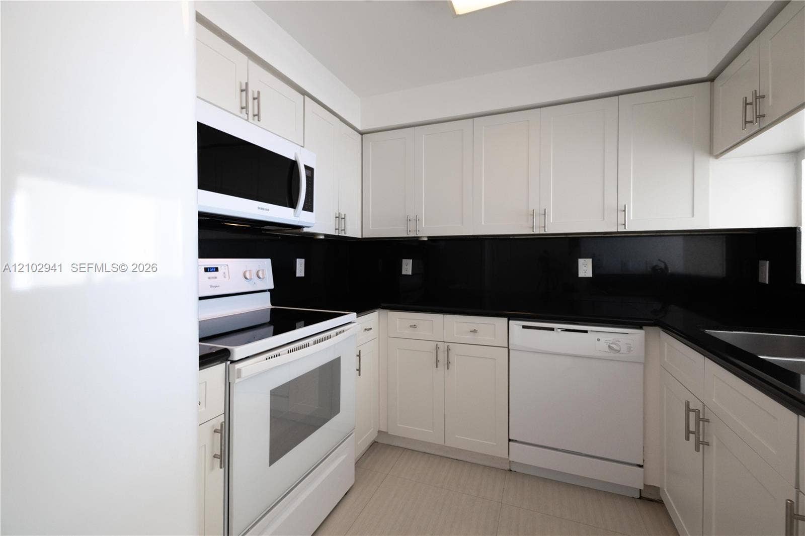 2475 Brickell Avenue, #1807, Miami, FL 33129 Kitchen featuring white shaker-style cabinetry, black stone countertops, matching black backsplash, and white appliances including a built-in microwave and dishwasher
