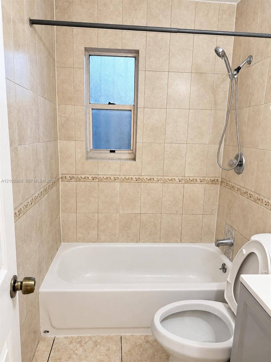 Bathroom featuring floor-to-ceiling neutral ceramic tile, decorative tile inlay, integrated shower and tub enclosure, handheld shower fixture, and privacy window