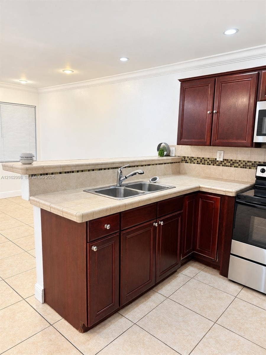 Kitchen featuring dark wood-finish cabinetry, tile countertops, decorative glass-tile backsplash, stainless steel sink with pull-down faucet, and neutral tile flooring