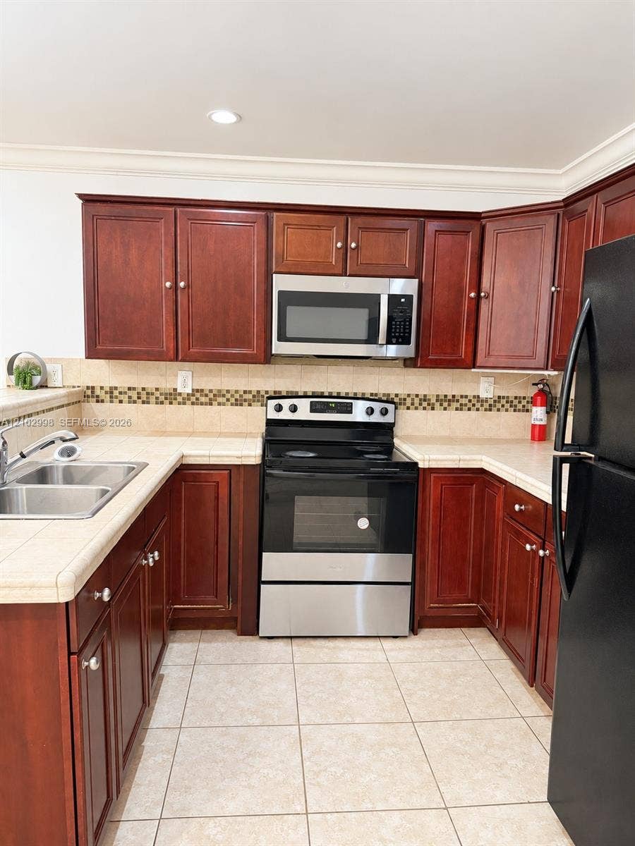 Kitchen featuring cherry-finish cabinetry, tile countertops, glass mosaic backsplash, stainless steel microwave, and tile flooring