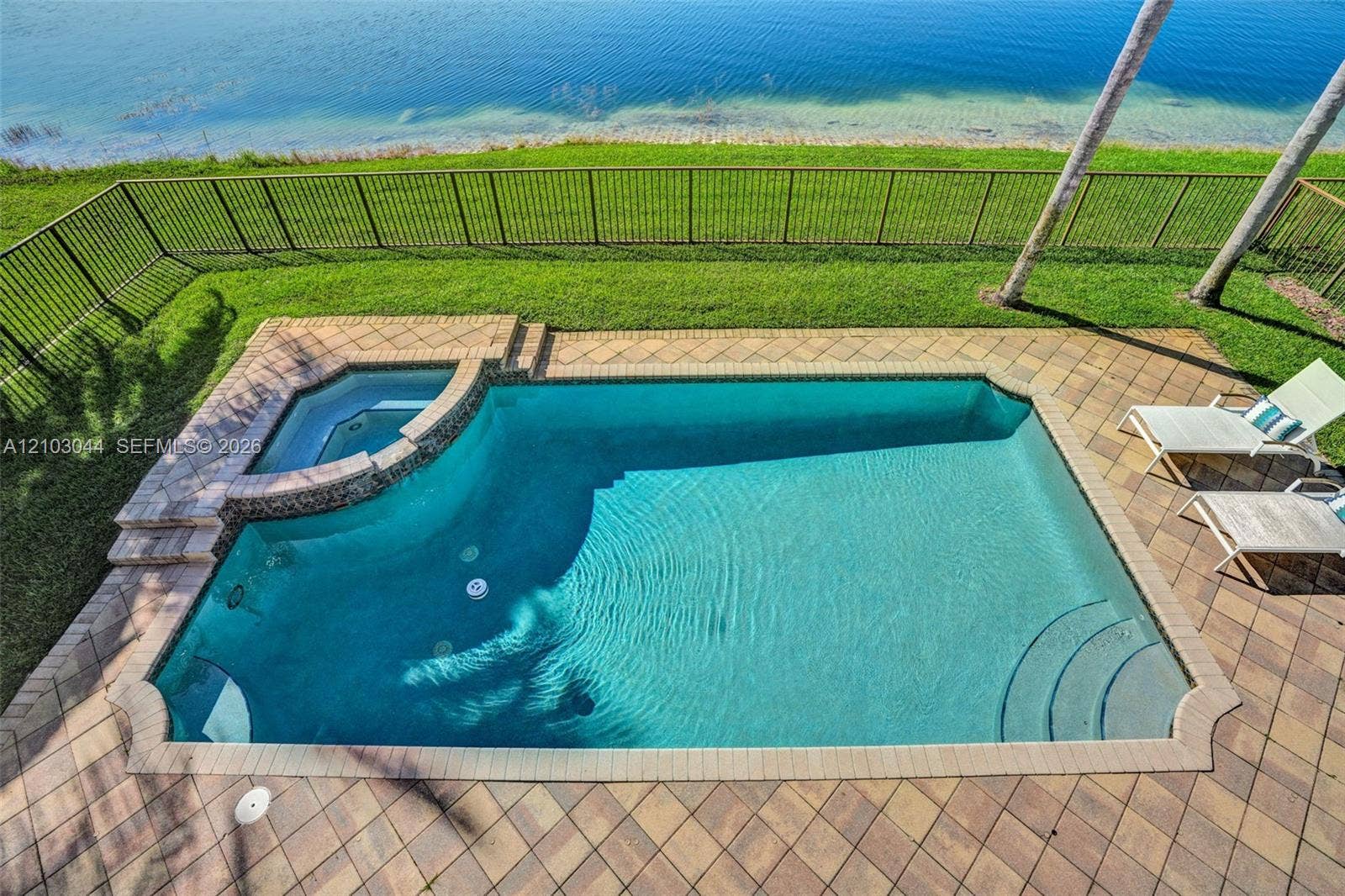In-ground pool with an integrated spa, paver deck, metal fencing, and grass lawn bordering the water