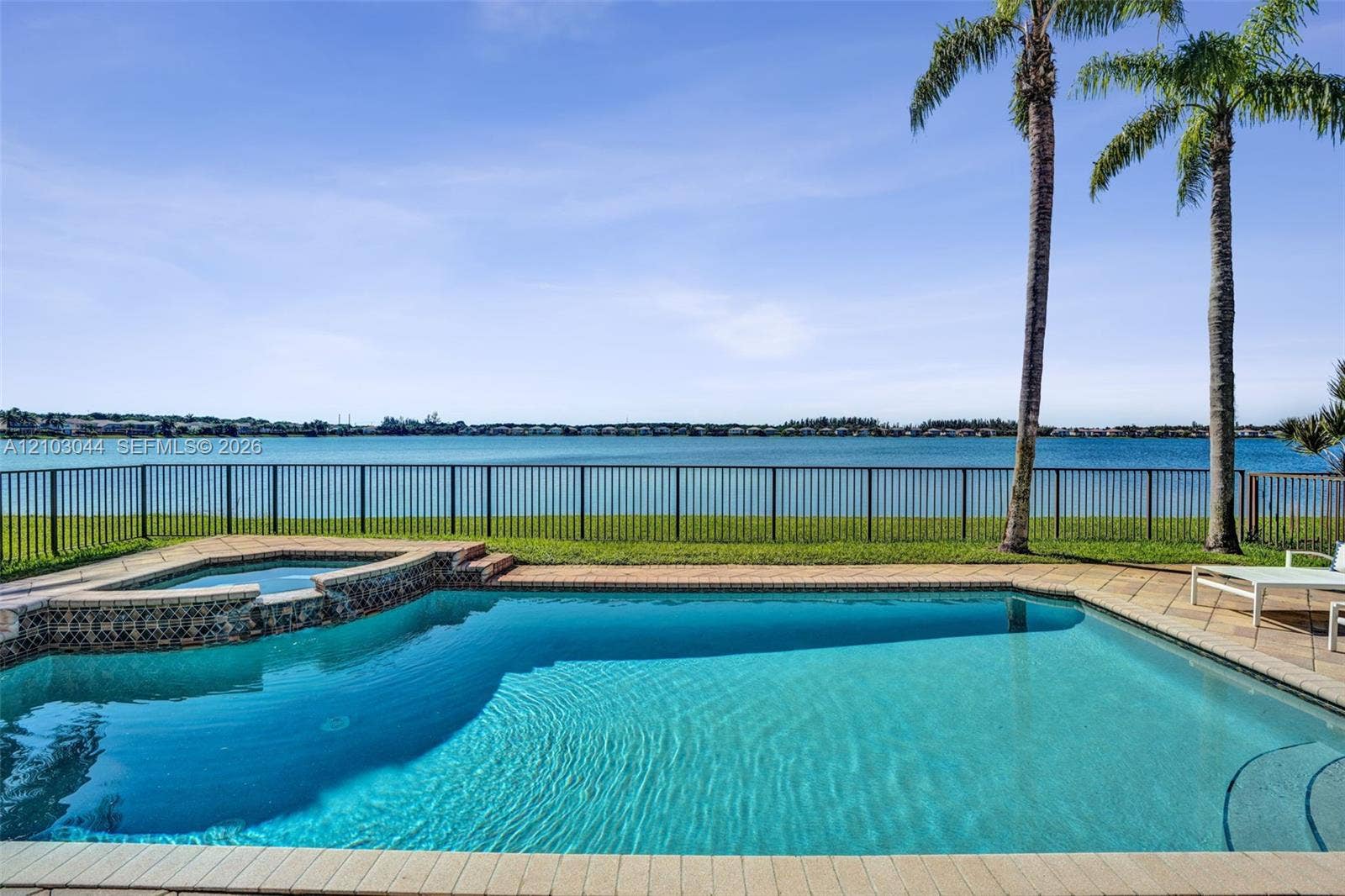 In-ground swimming pool with an integrated spa, stone-paver decking, and metal perimeter fencing overlooking a lake with mature palm trees