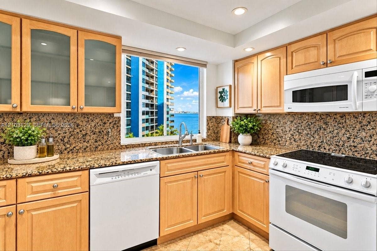 1627 Brickell Avenue, #2005, Miami, FL 33129 Kitchen featuring natural wood cabinetry with glass-front uppers, stone countertops, full-height stone backsplash, double-basin stainless steel sink, and white appliances