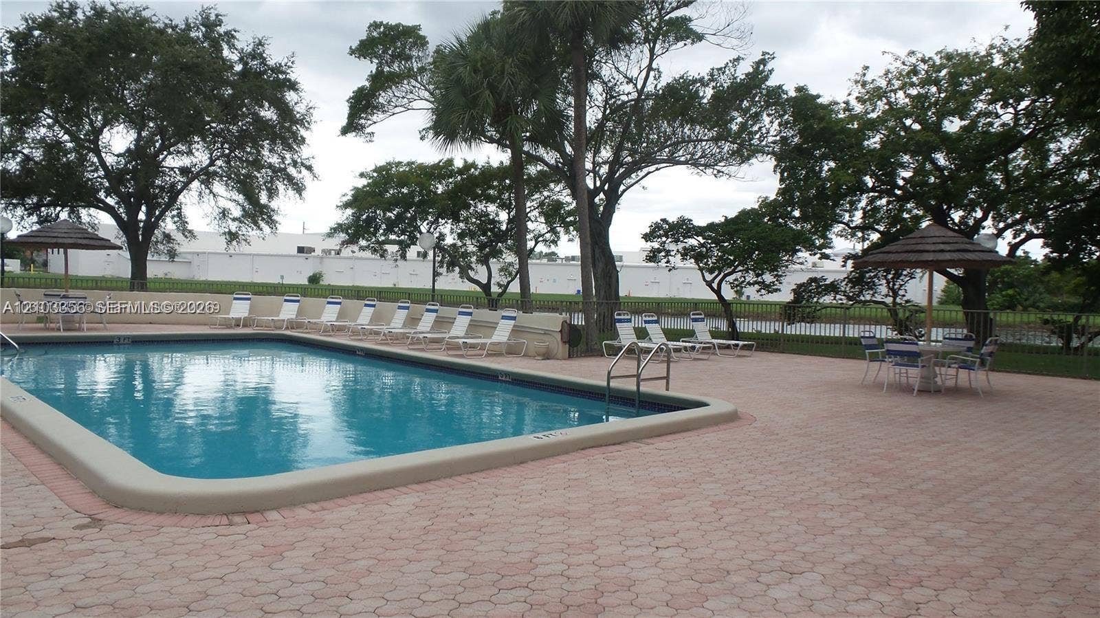 In-ground swimming pool featuring a concrete deck with brick-paver border, metal perimeter fencing, and mature trees