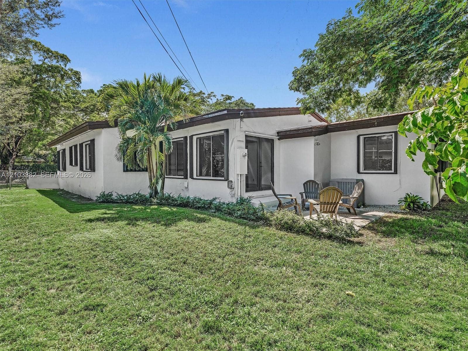 1545 Mantua Avenue, Coral Gables, Coral Gables, FL 33146 Exterior featuring white stucco siding, dark-trimmed windows, a concrete patio, and a fenced grass lawn with mature trees and palm foliage. House for rent in Coral Gables. House for rent listed by Realtor.