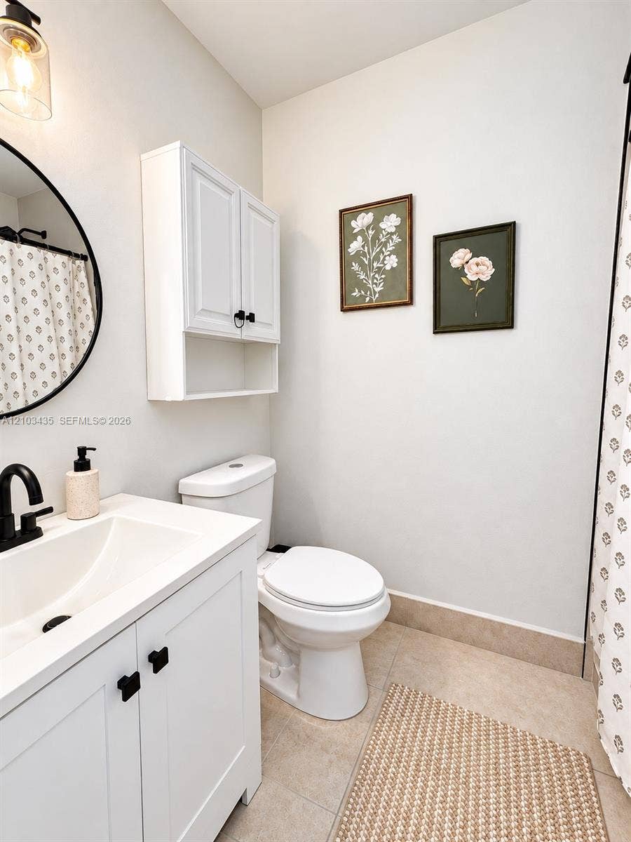 Miami, FL 33177 White vanity with a rectangular sink, matte black faucet, wall-mounted storage cabinet, circular mirror, and tile flooring