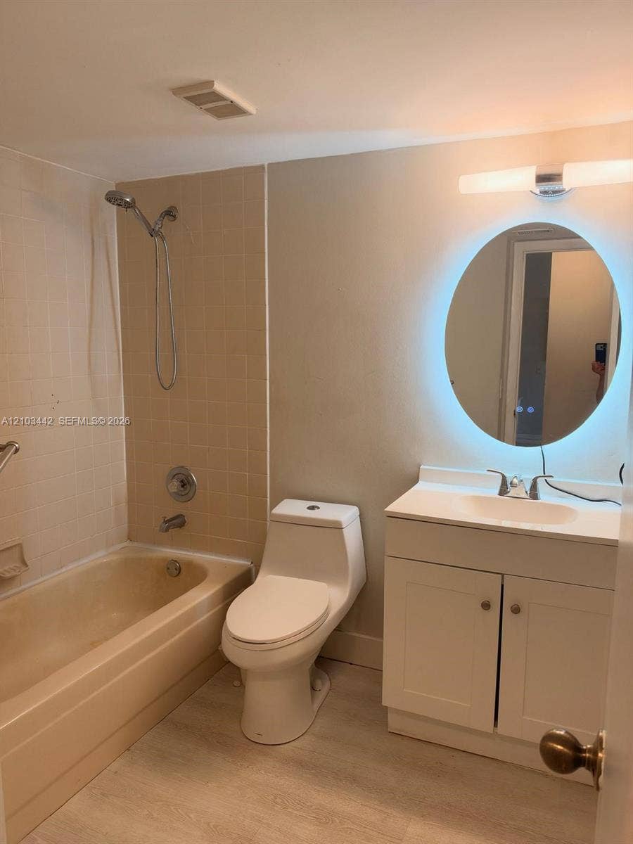 8721 Wiles Road, #303, Coral Springs, FL 33067 Bathroom featuring a white vanity cabinet with a solid-surface countertop, an integrated sink, an illuminated oval mirror, a porcelain commode, and a tub-shower combination with ceramic tile surround