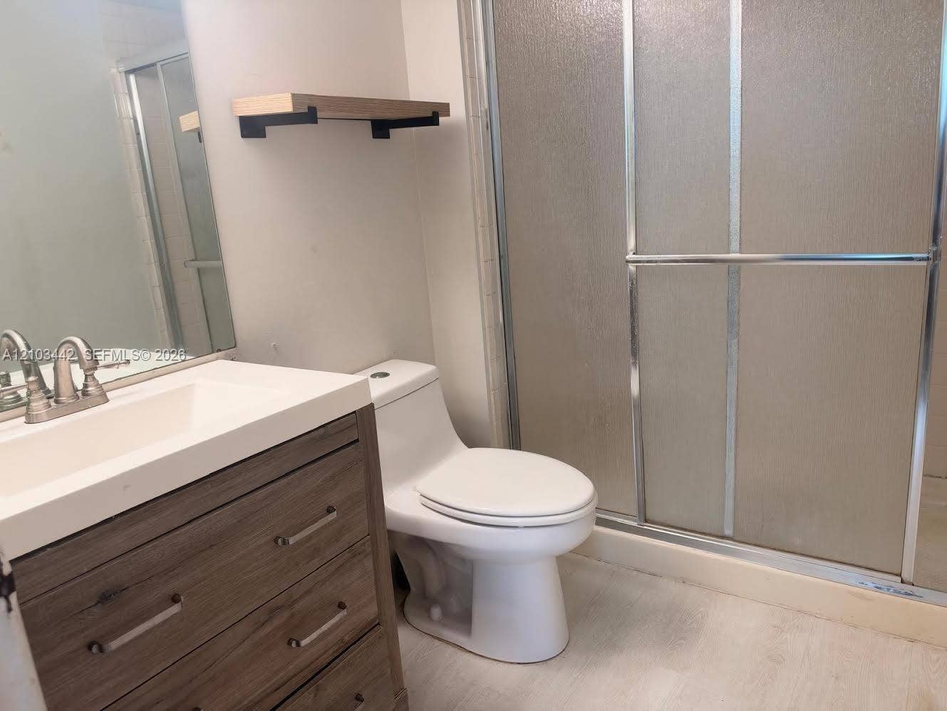8721 Wiles Road, #303, Coral Springs, FL 33067 Bathroom featuring a vanity with a wood-finish cabinet and integrated sink, a large wall-mounted mirror, a built-in wood shelving unit, and a walk-in shower with a glass enclosure
